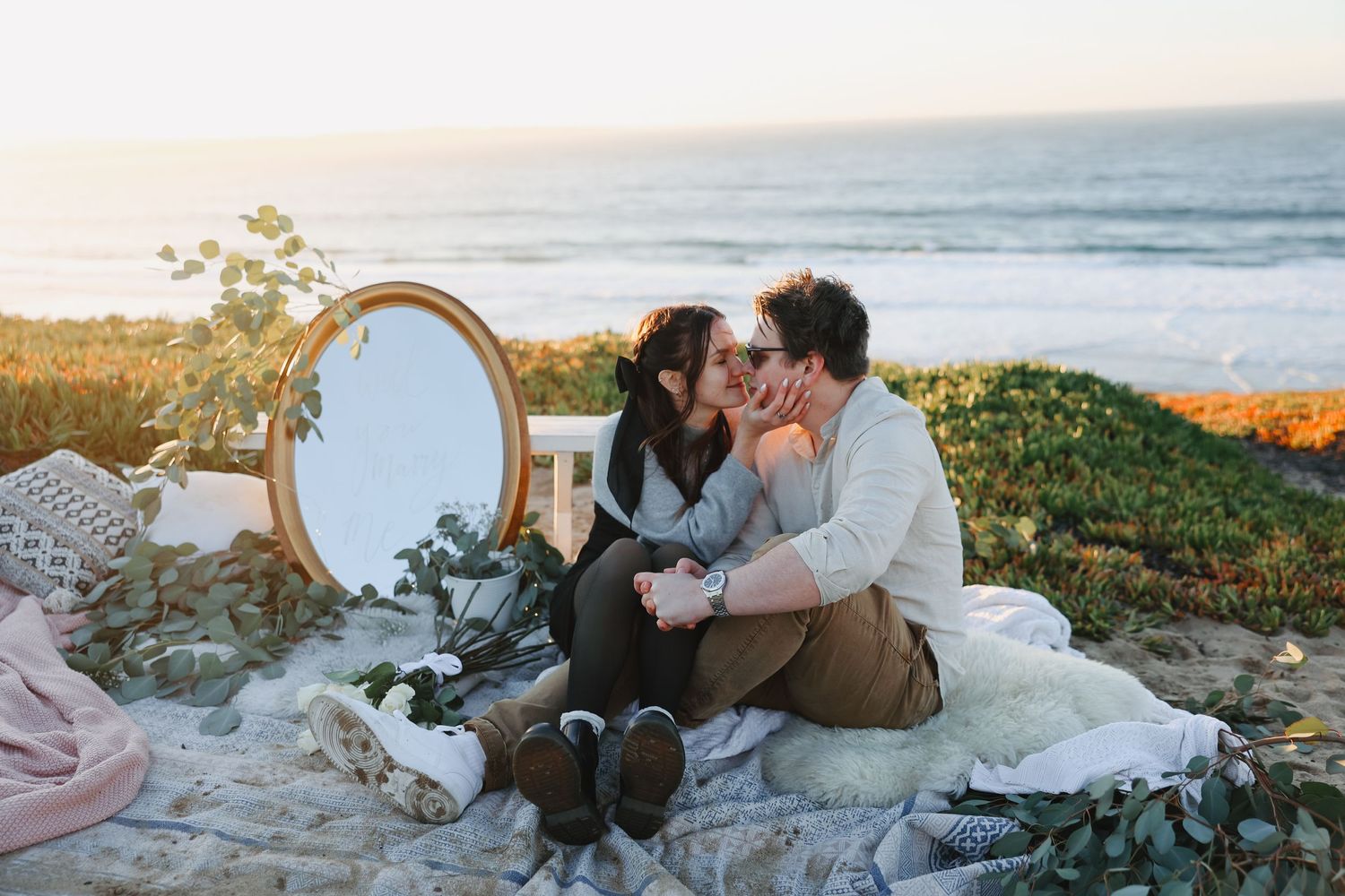 Couple enjoying romantic seaside picnic setup with vintage mirror and blanket at sunset.