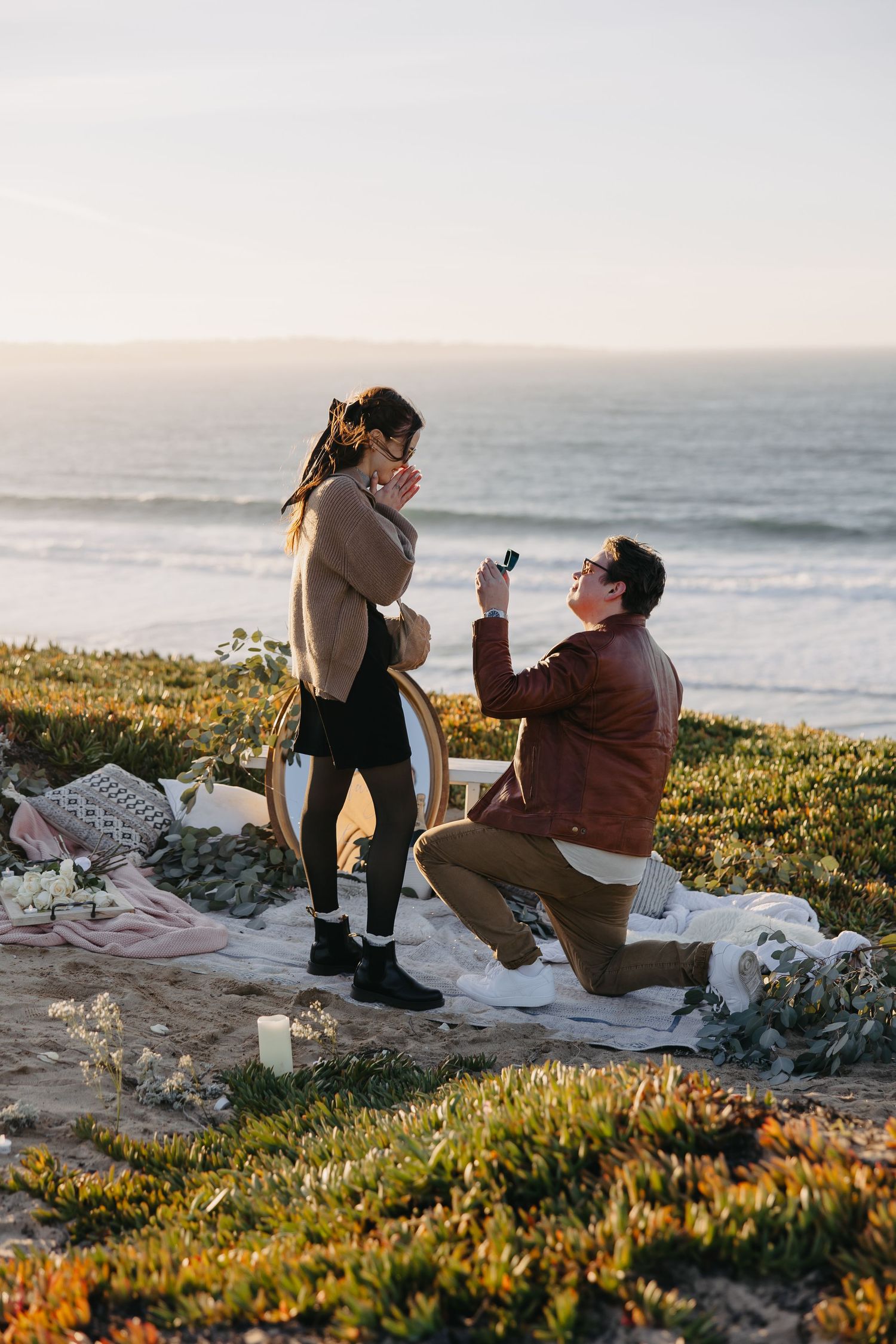 A romantic proposal scene at sunset on a coastal cliff with a picnic blanket and candles set up.