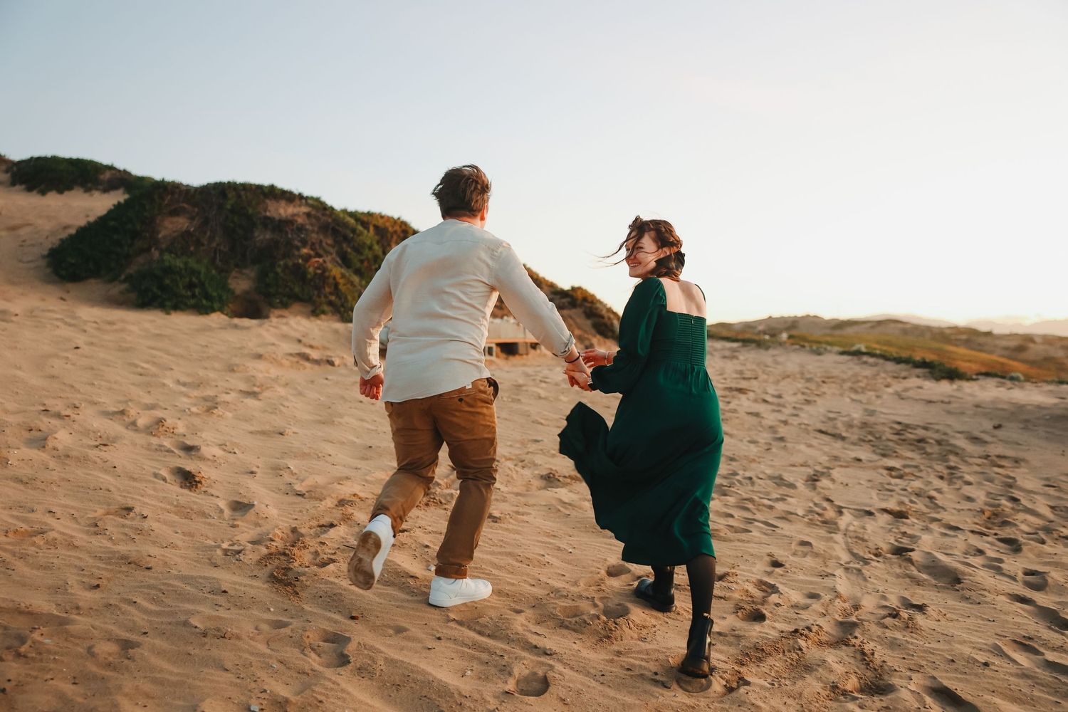 Playful couple runs hand in hand up sandy dunes during sunset photoshoot.