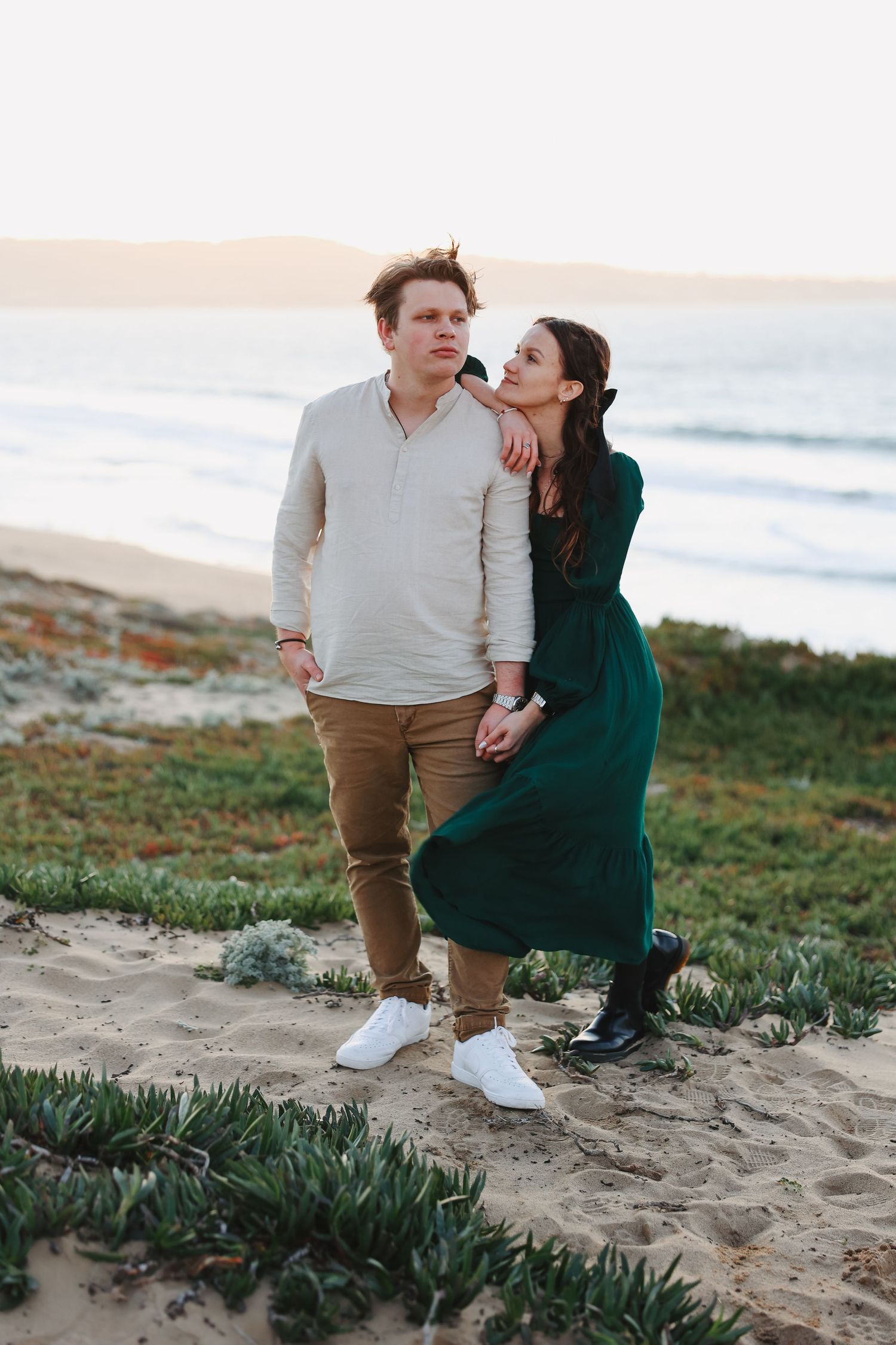 A couple in contrasting outfits poses together on a coastal path at sunset.