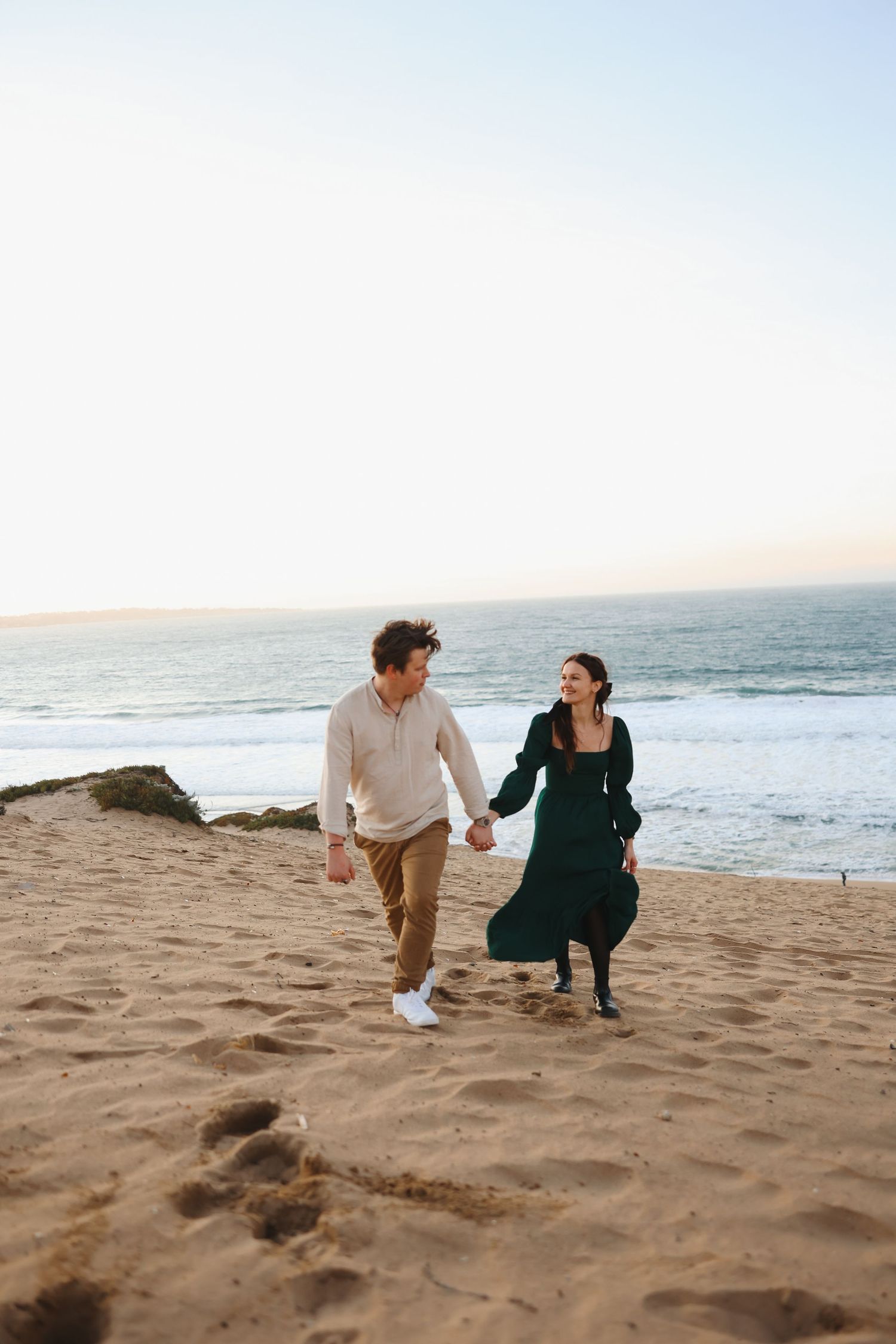 A couple holds hands while walking on a sandy beach at sunset wearing casual attire.