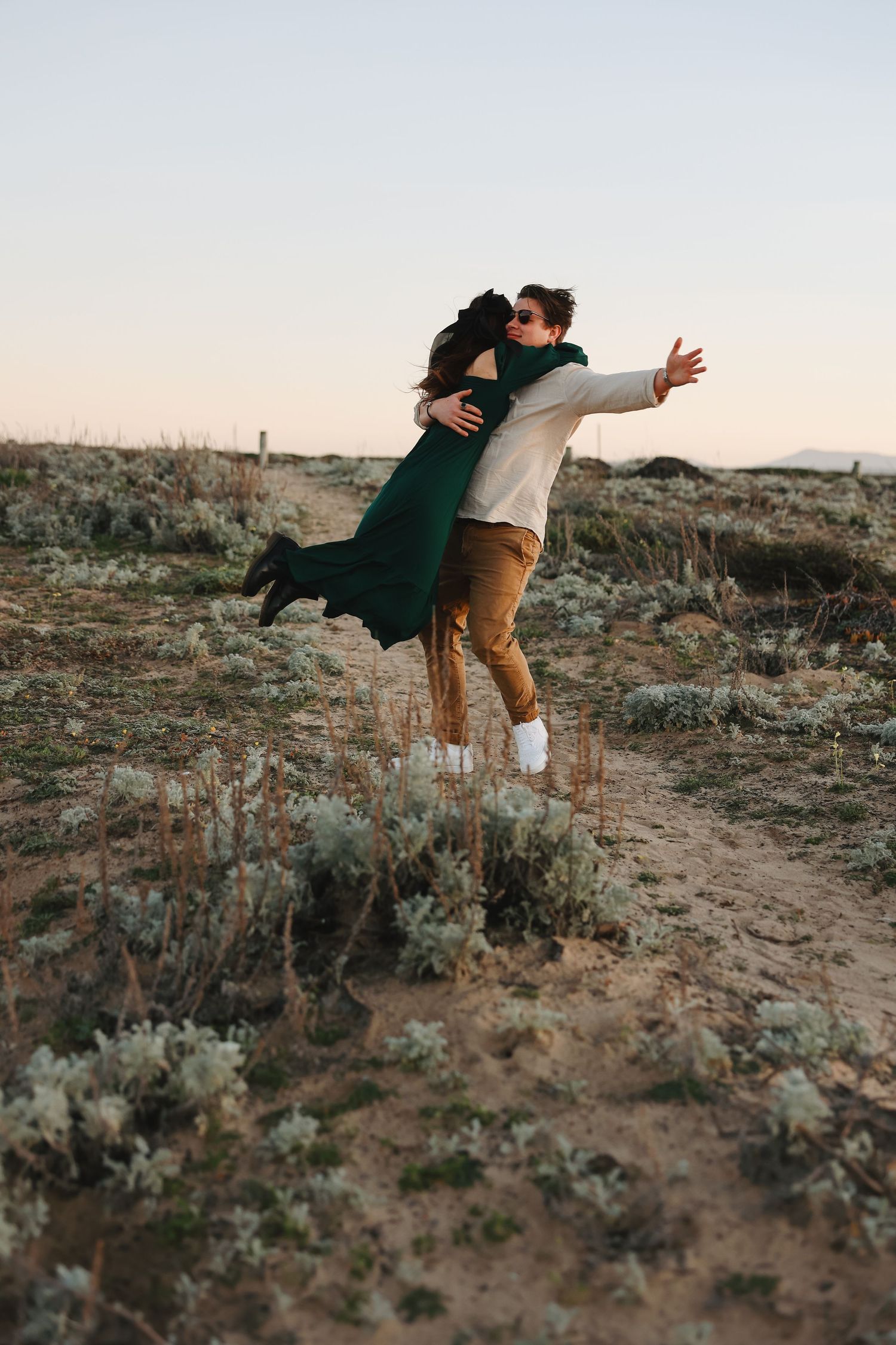 A couple shares a playful embrace while standing on a dirt trail surrounded by desert brush.