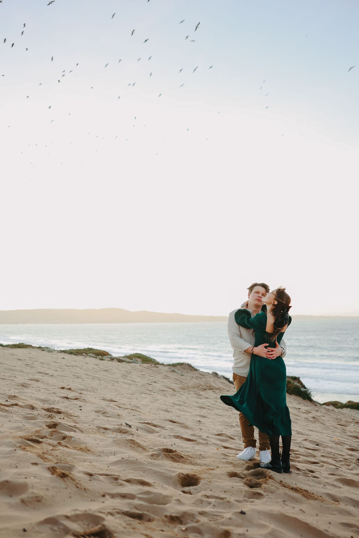 A romantic moment shared between two people on a beach with seabirds flying overhead.