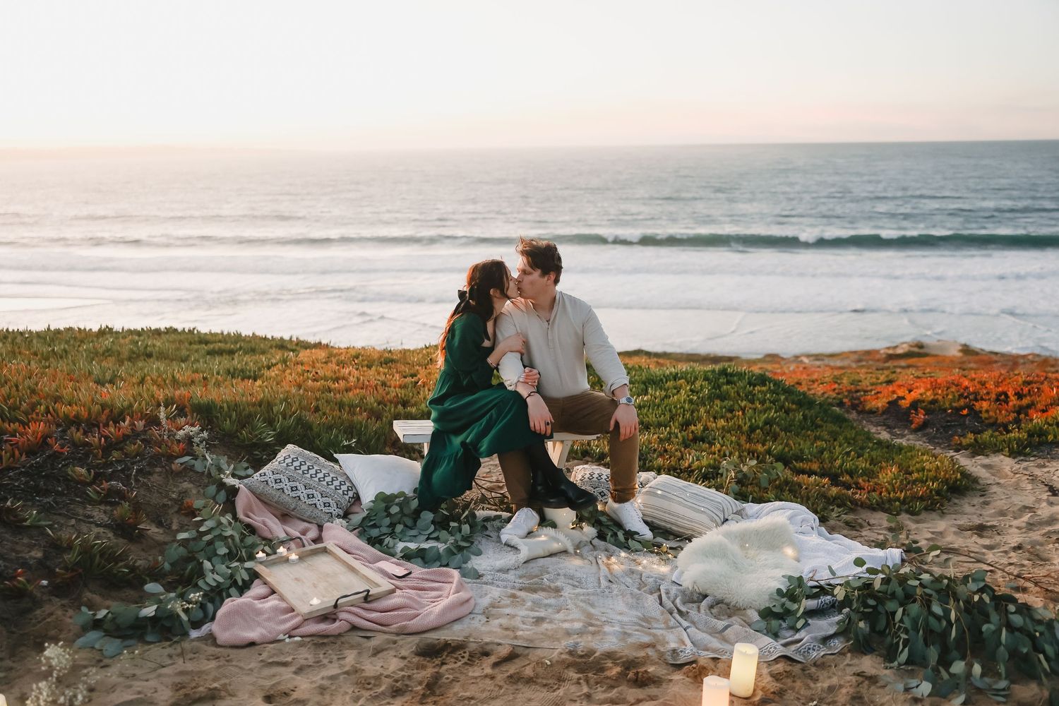 A cozy seaside picnic at golden hour with blankets spread out on grassy cliffs.