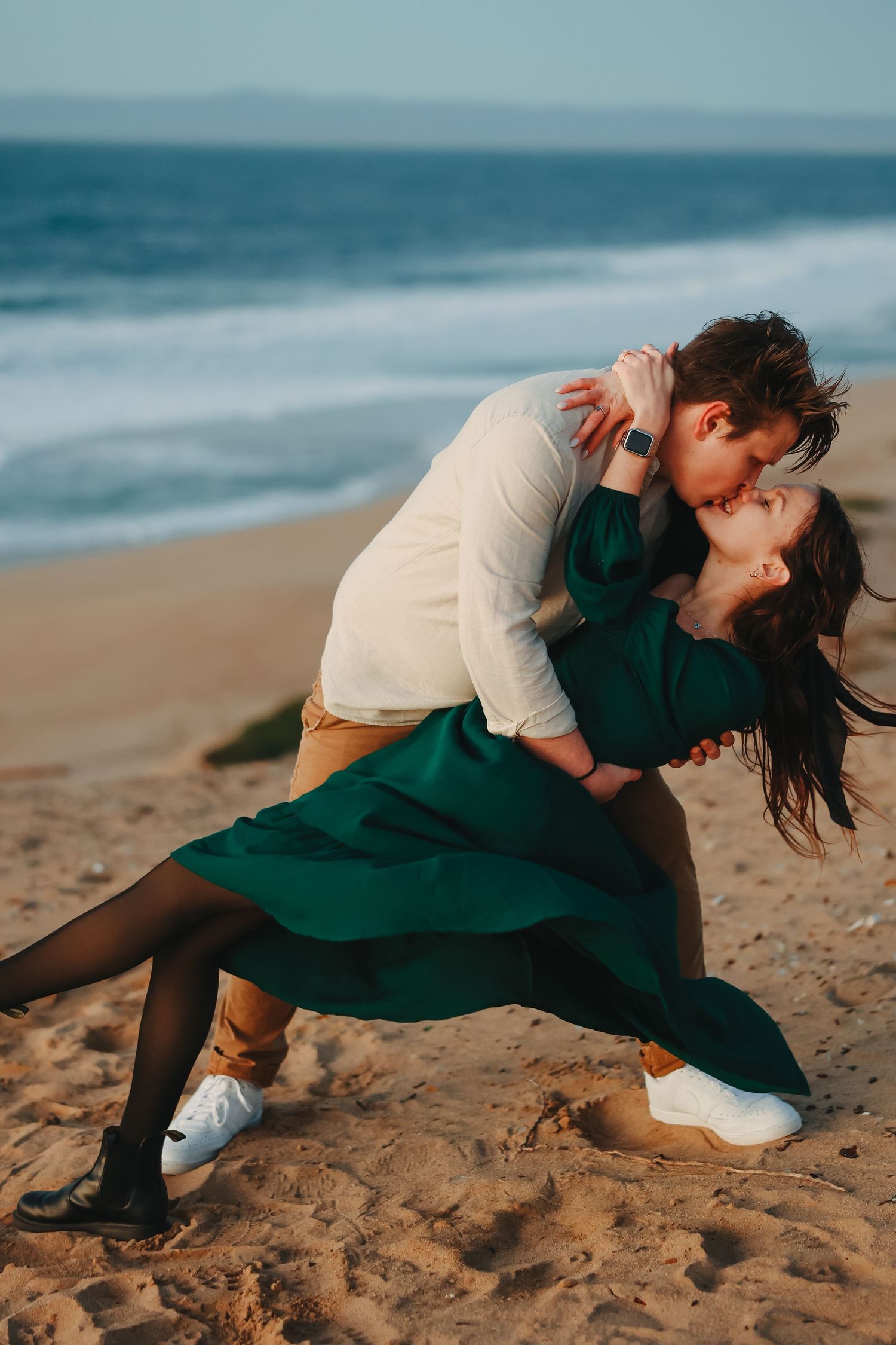 Couple performs classic dance dip pose on sandy beach with waves crashing behind them.
