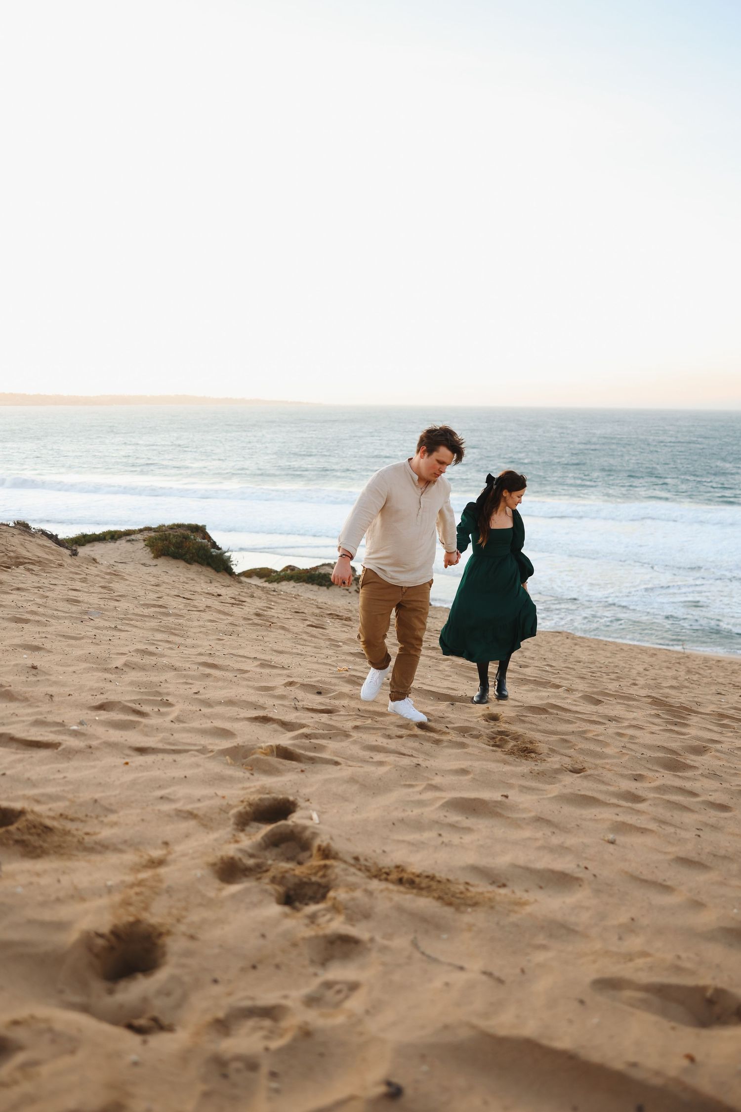 A couple descends a sandy slope together near the ocean during golden hour.