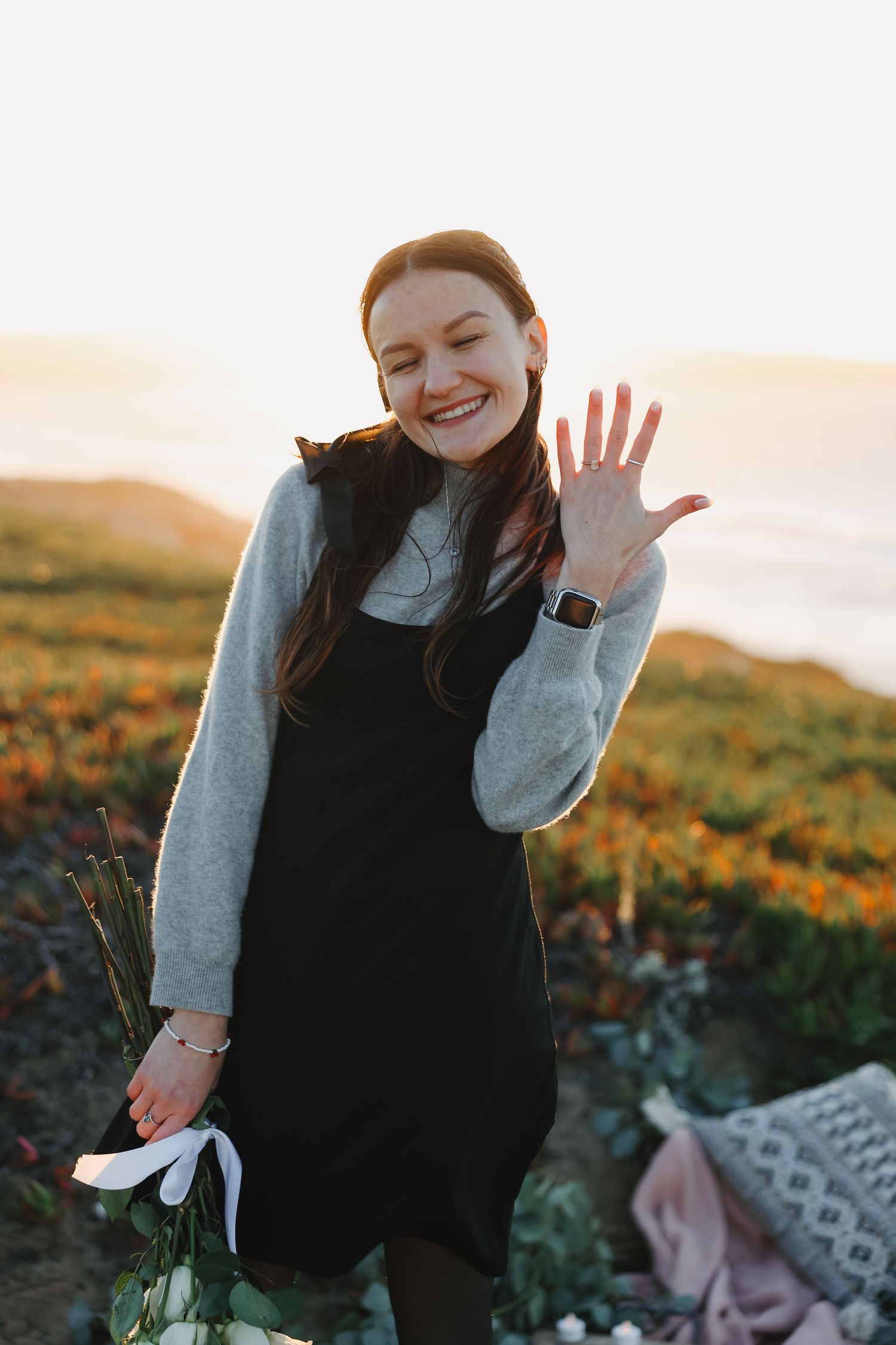 Woman showing engagement ring while smiling on coastal cliff at sunset.
