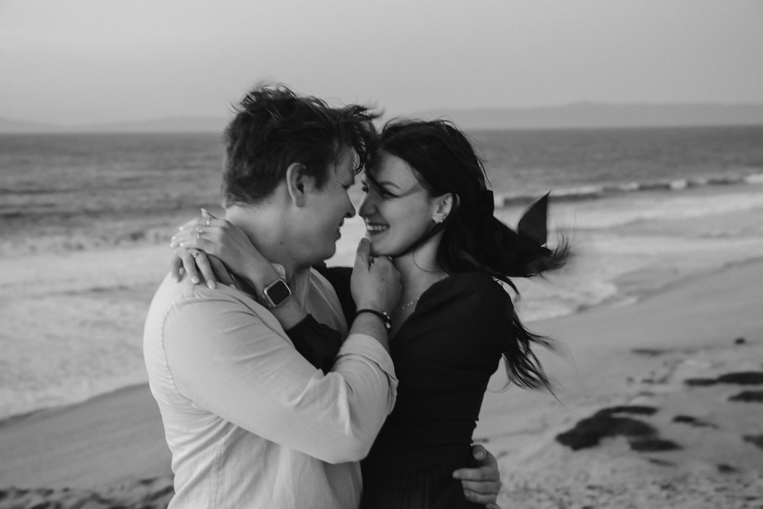 A black and white beach portrait showing an intimate moment between two people with ocean waves behind.