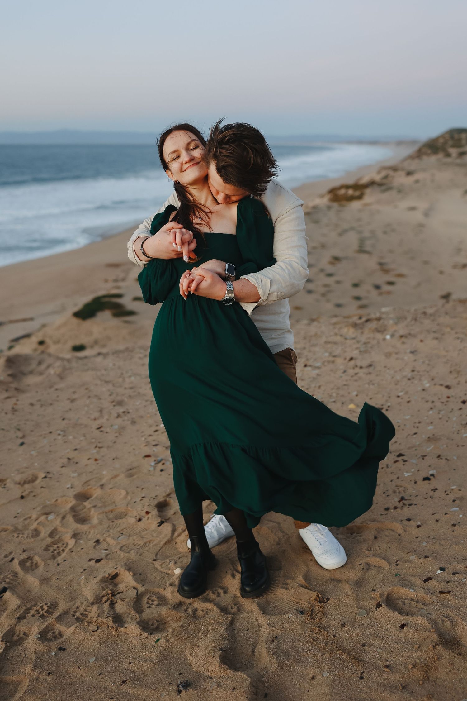 A romantic embrace on the beach with flowing green dress and casual attire at sunset.