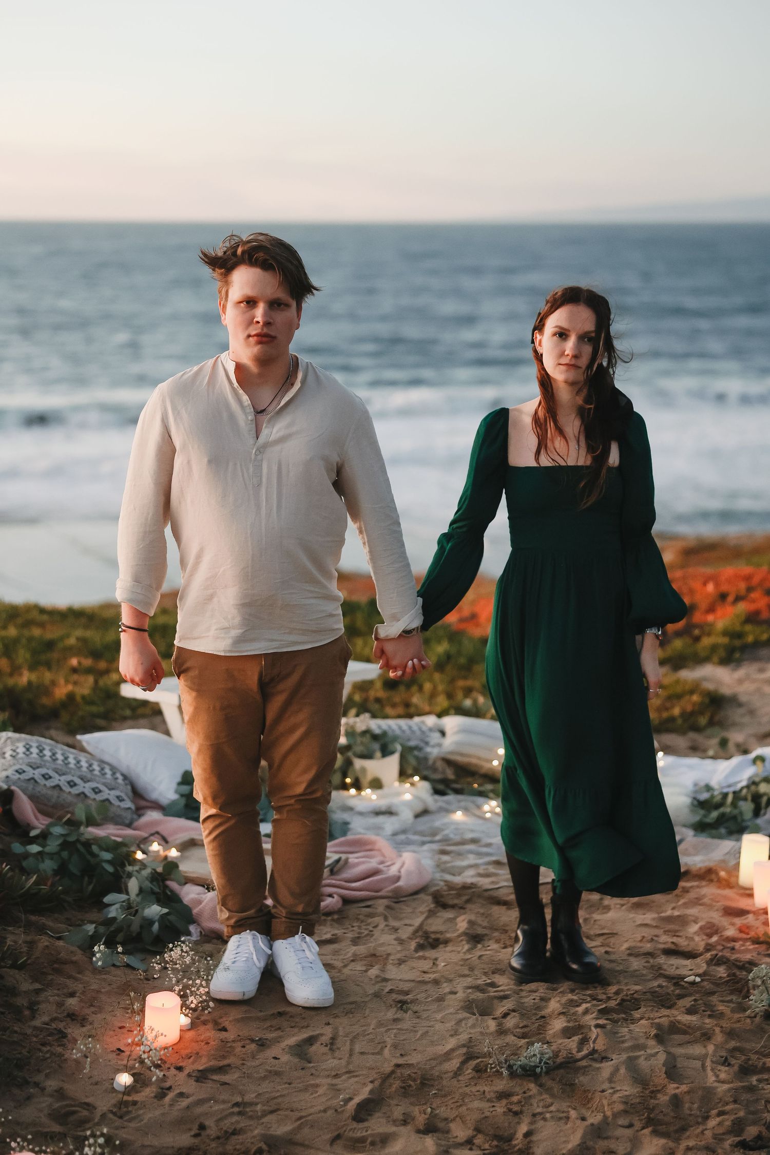 A couple holds hands on a beach at sunset with candles glowing in the foreground.