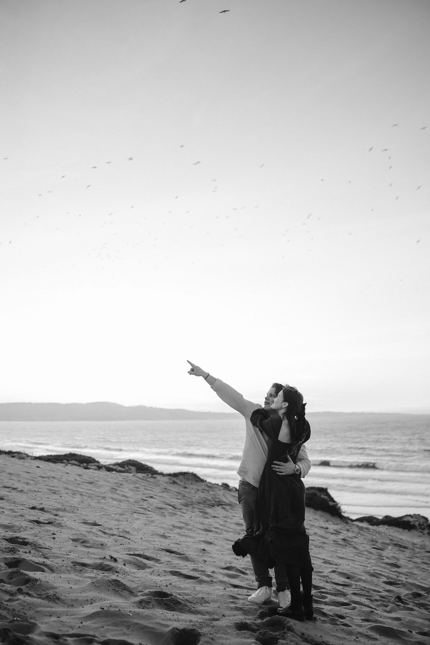 A couple points at birds while sharing a moment on a beach in black and white photography.