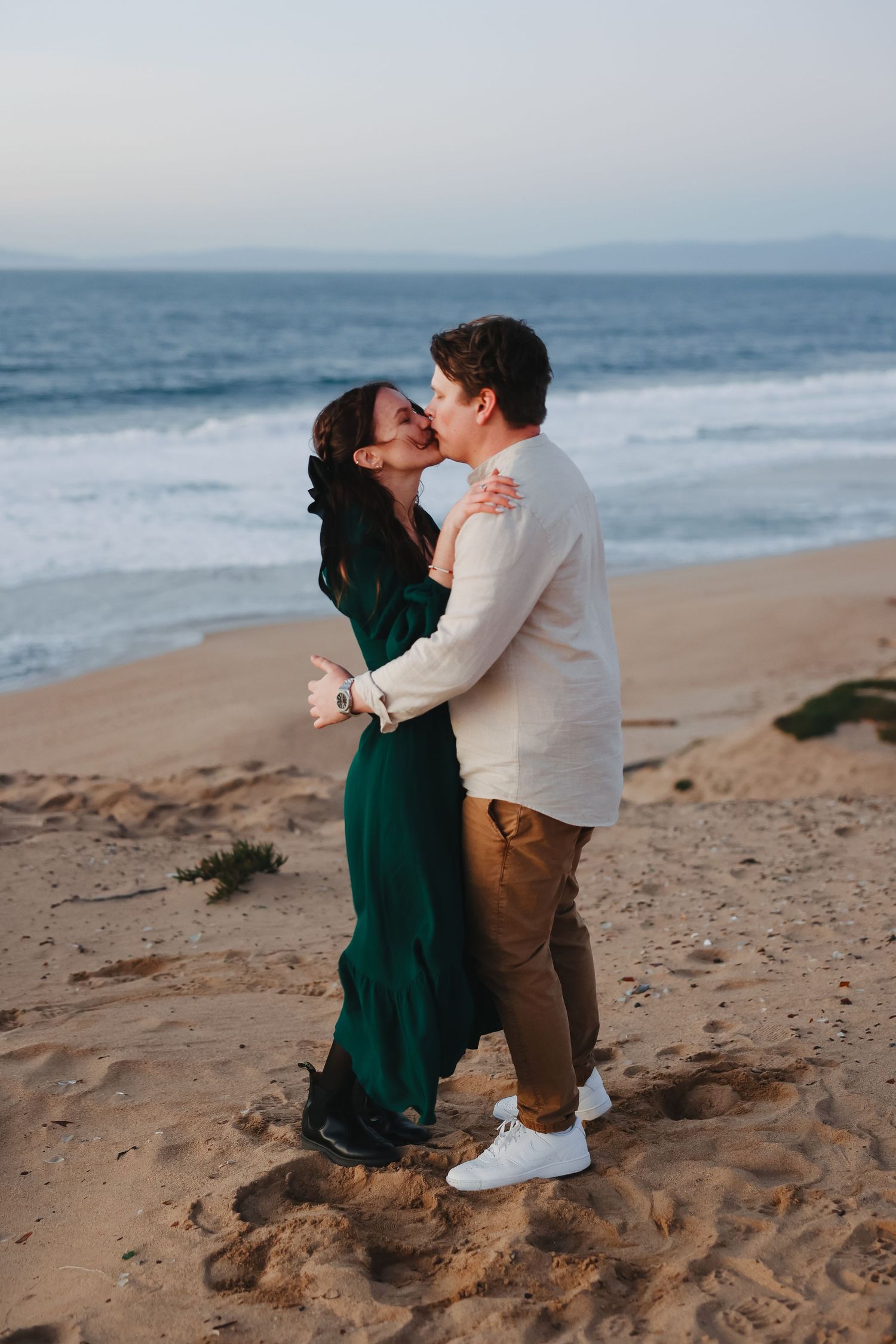 A romantic couple embraces on a sandy beach with waves crashing in the background at dusk.