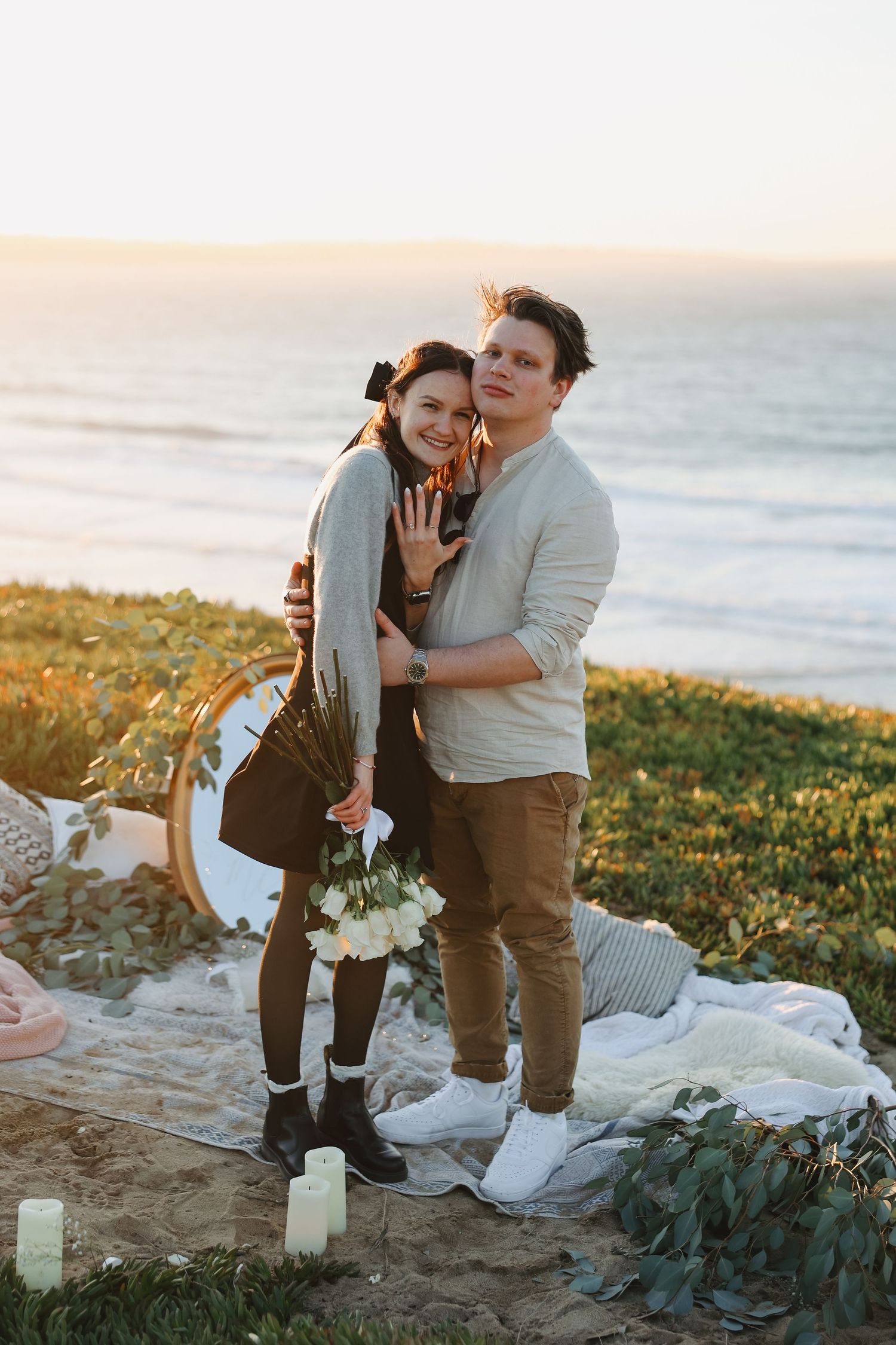 Happy couple embracing on scenic cliff after romantic proposal with flowers and candles.