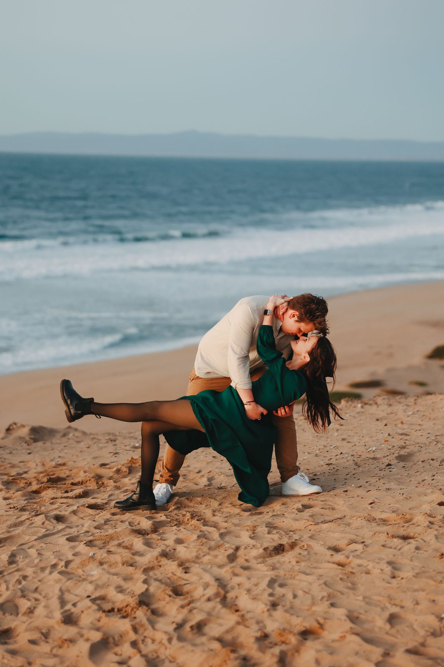 Couple performs dramatic dance dip on beach with emerald dress flowing in ocean breeze.