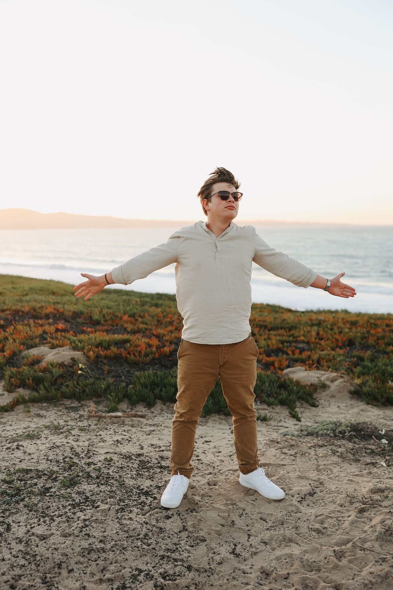 Person in white sweater and khakis standing with arms spread on coastal cliff at sunset.