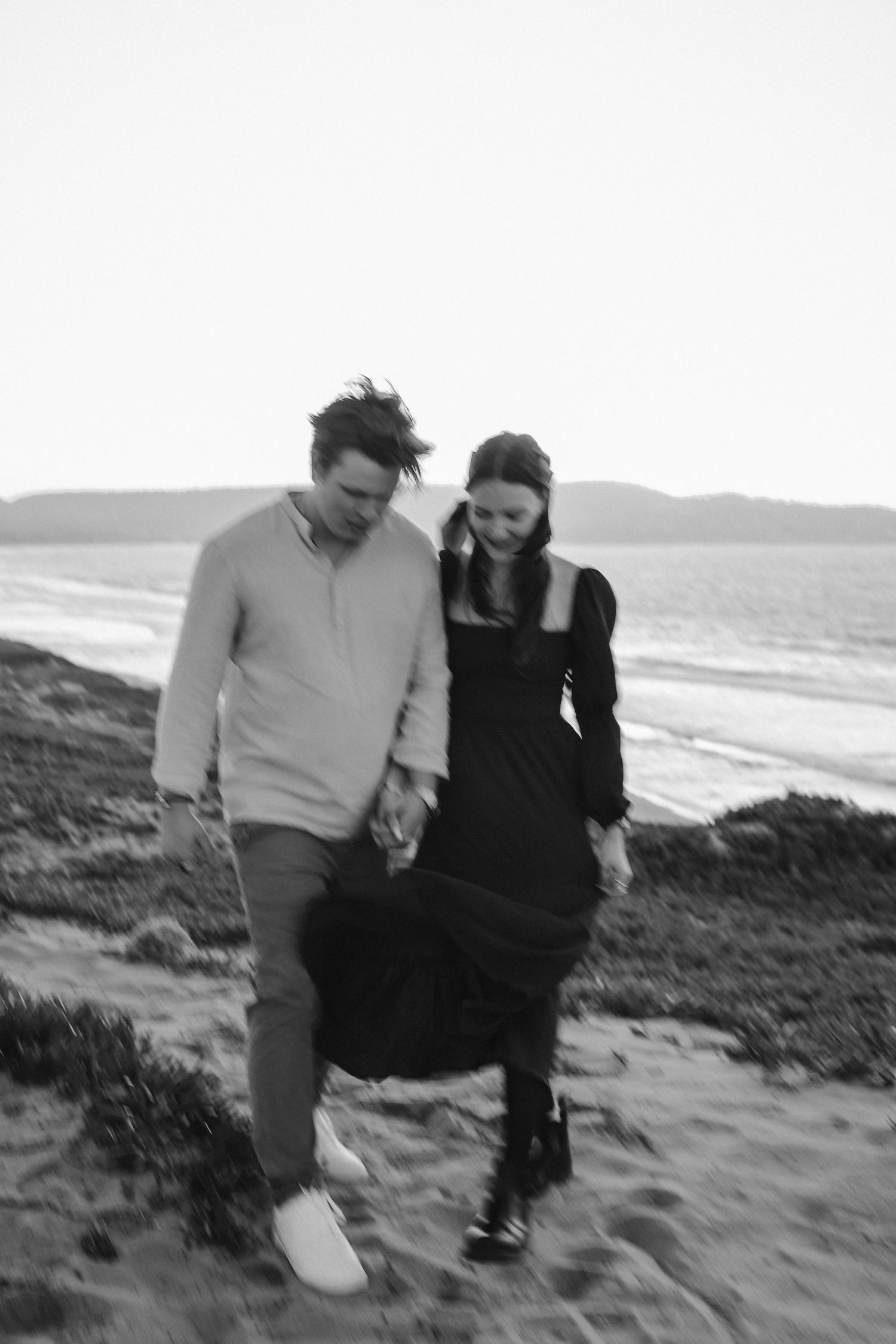 A couple holding hands walks together on a sandy beach path with ocean waves in the background.