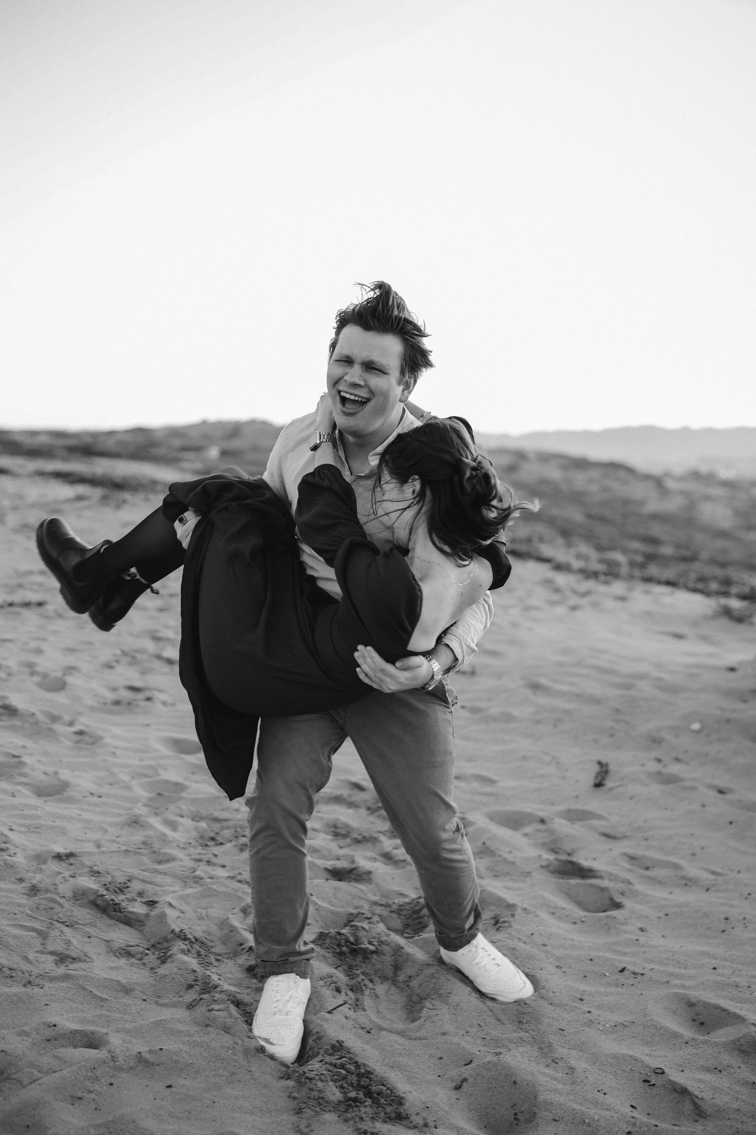 A black and white photo shows a couple sharing a playful moment with one being carried on the beach.