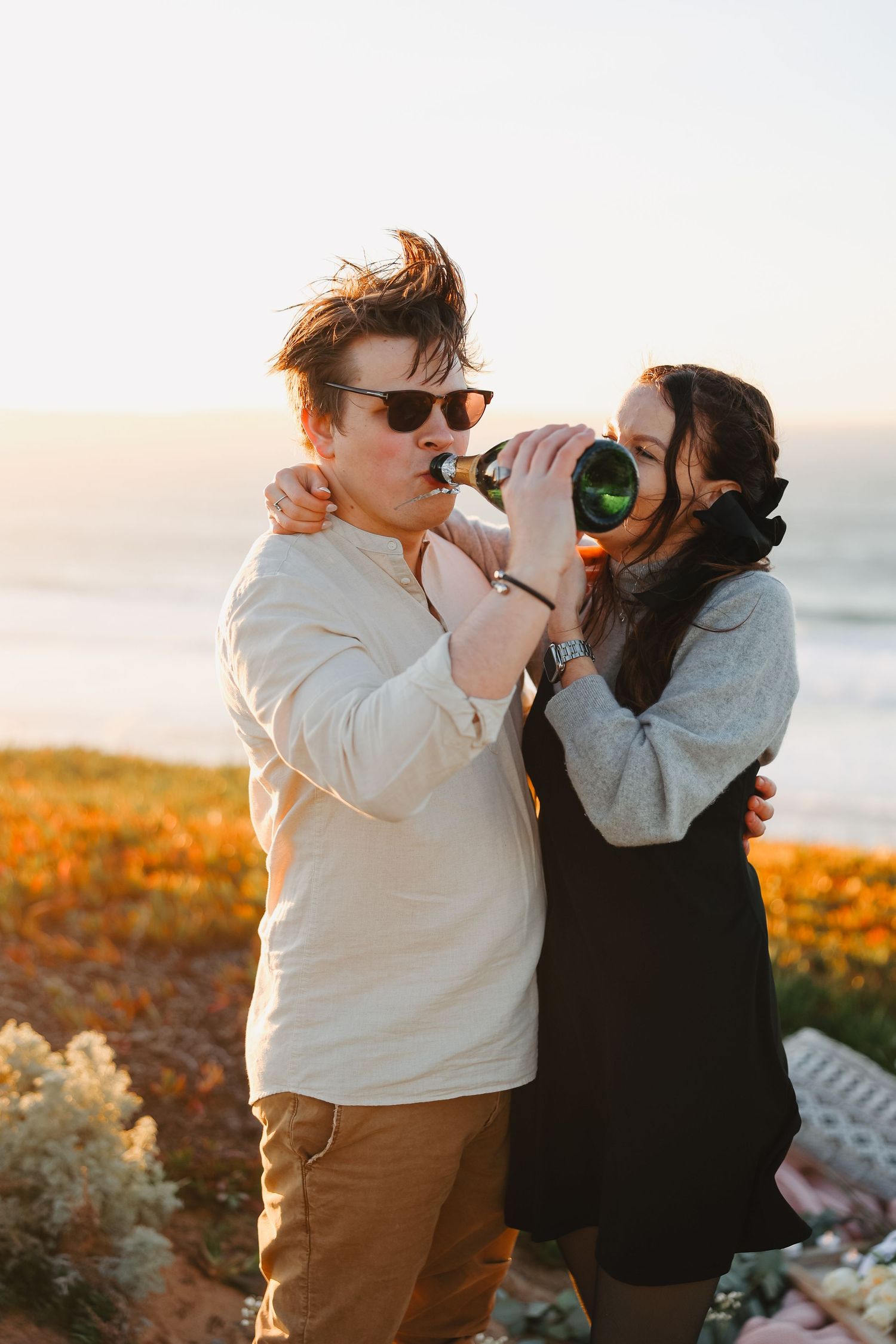 Playful couple drinking from bottles during sunset picnic by the ocean.