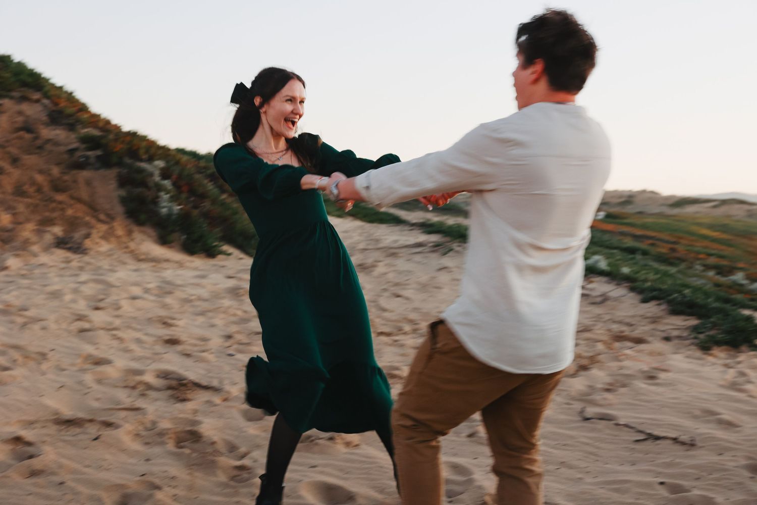 A couple dances and spins together on a sandy beach with cliffs in the background.