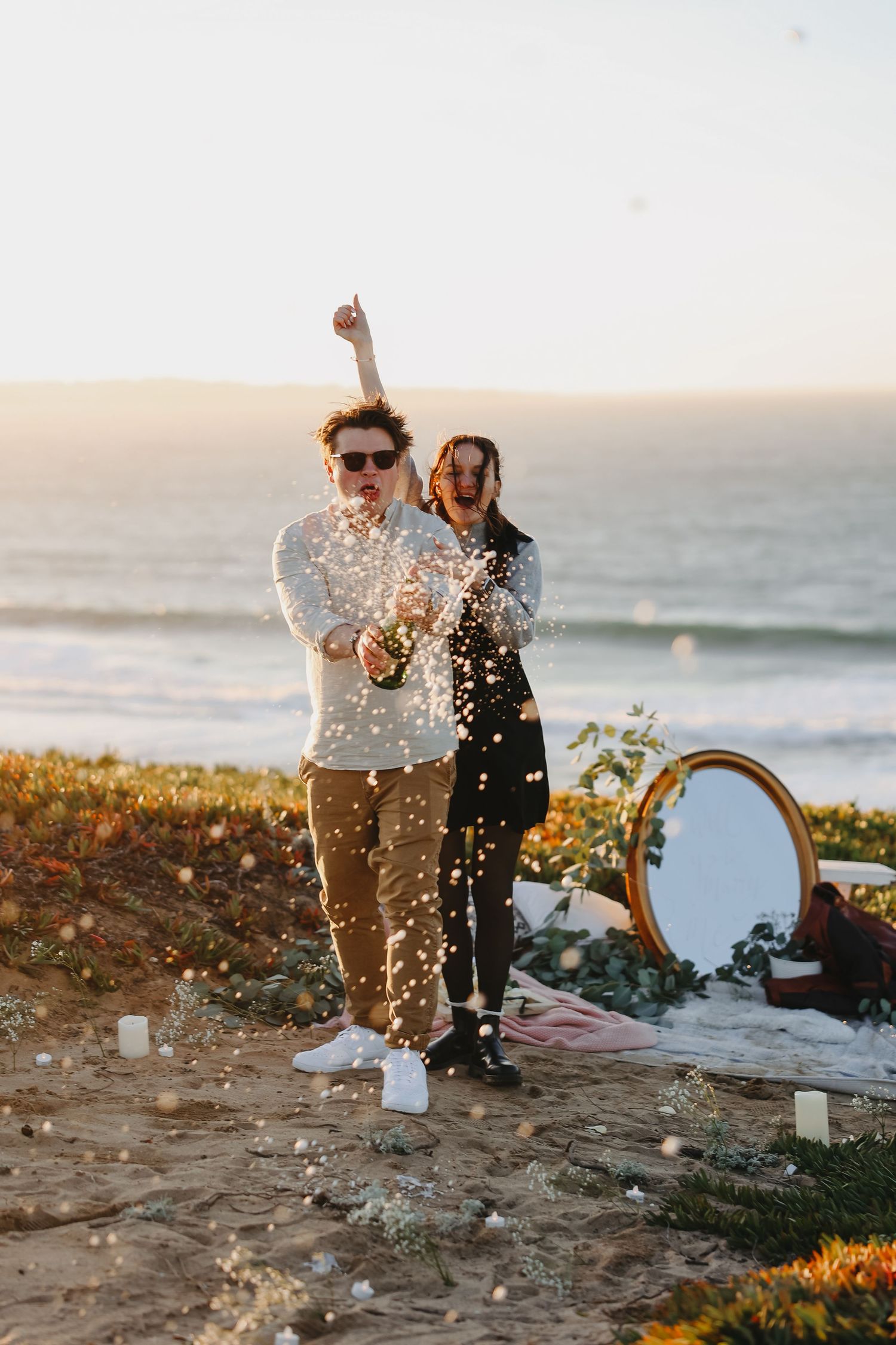 A couple celebrating with champagne by the ocean as the sun sets behind them.