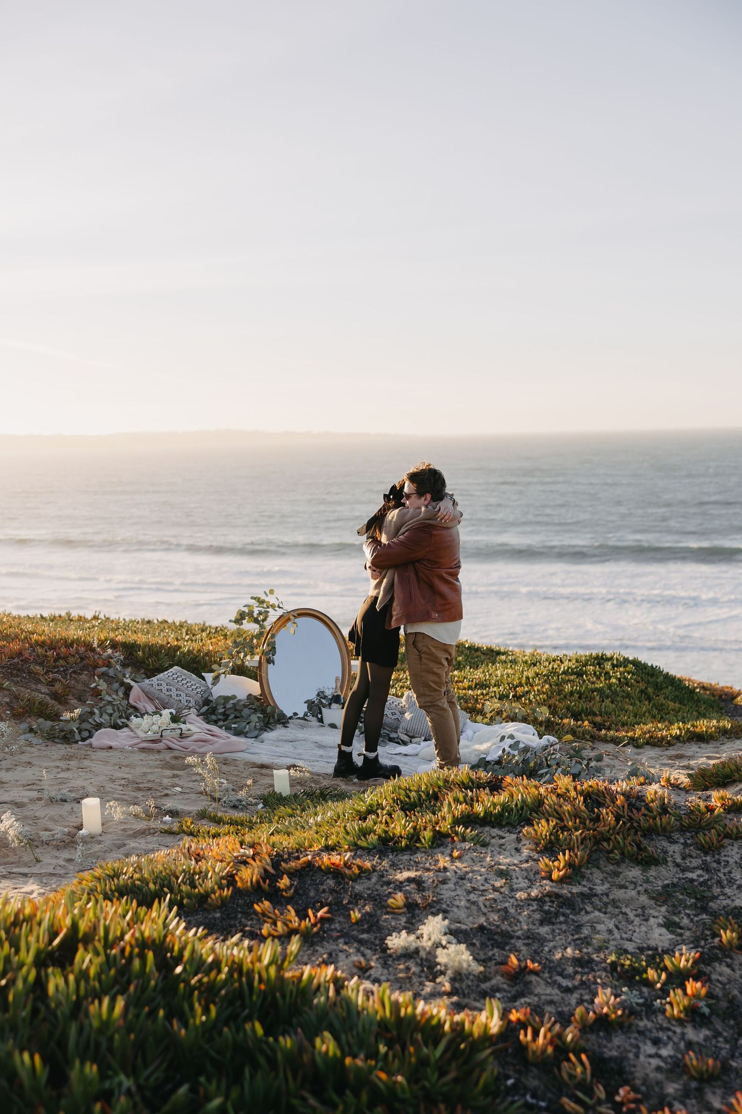 A couple embracing during a seaside proposal with decorative elements and golden hour lighting.