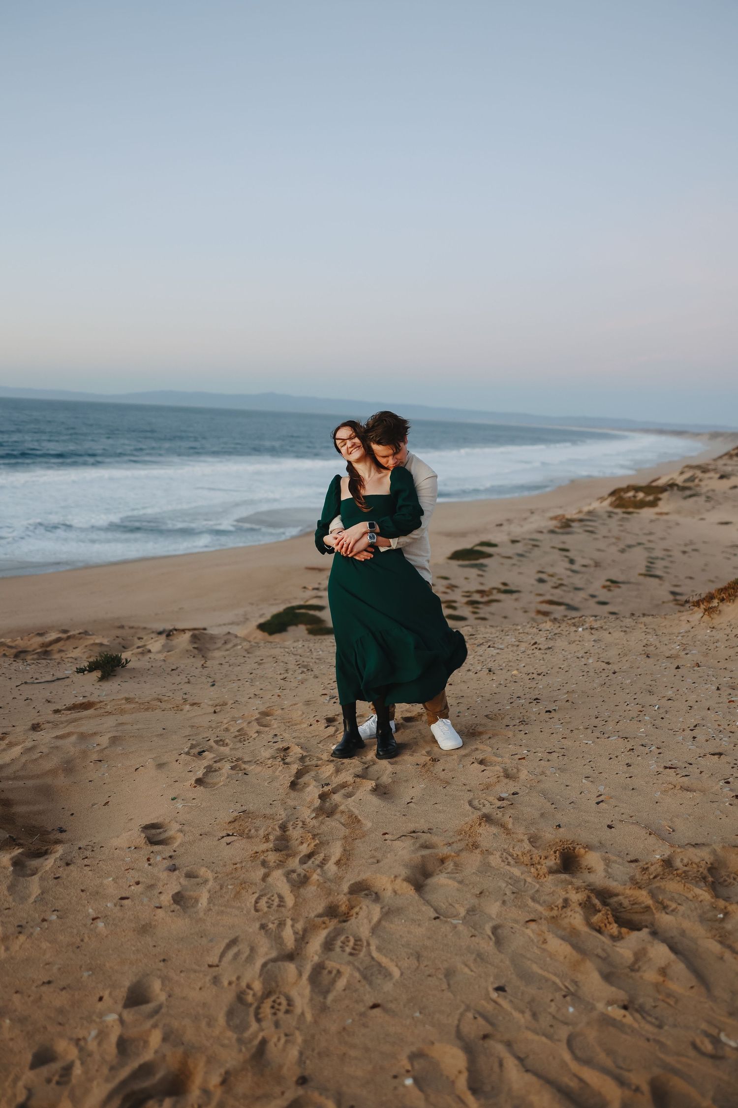 Couple in elegant attire embraces while standing on scenic coastal cliffs overlooking the ocean.