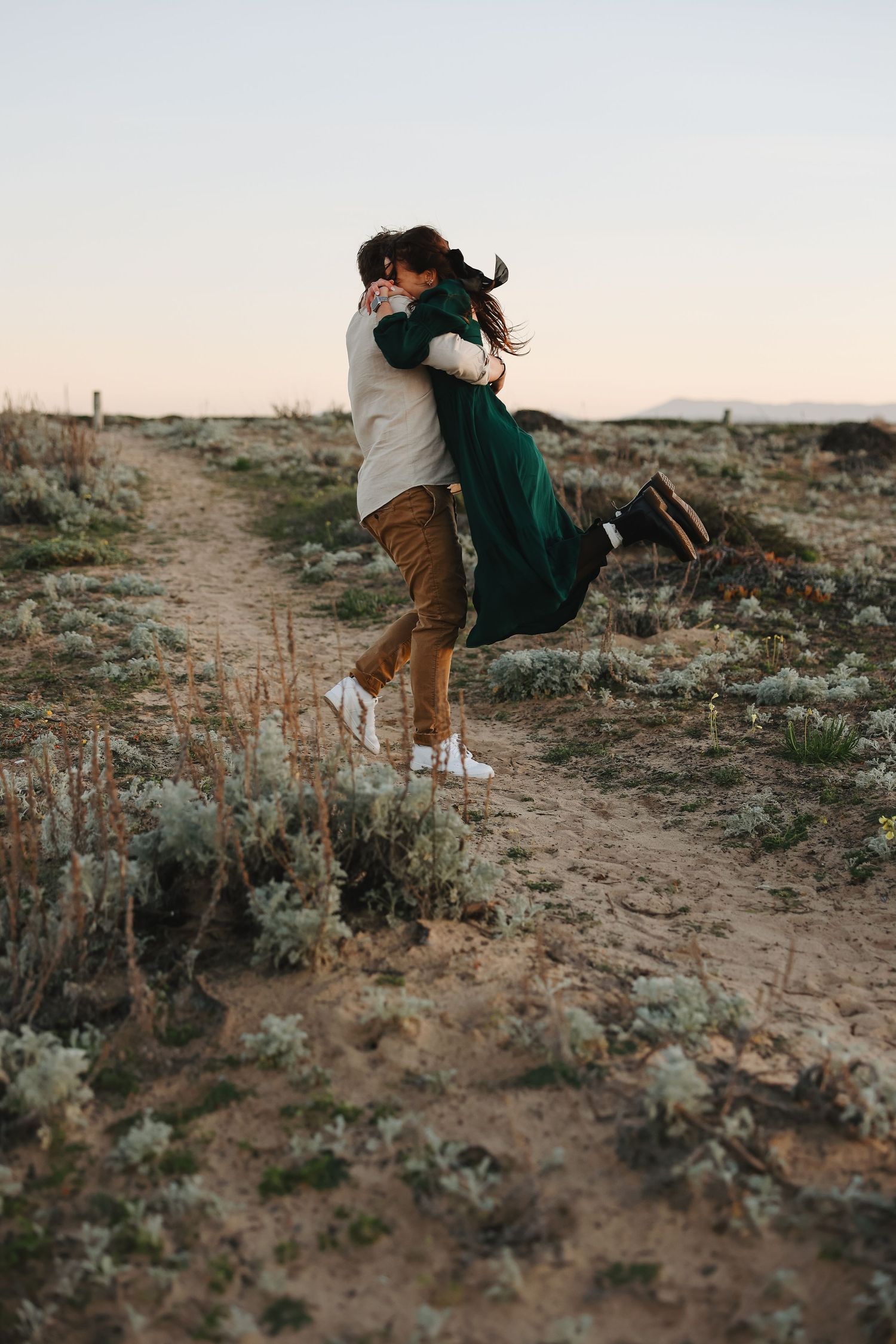 A romantic couple embrace and spin joyfully on a desert path during sunset.