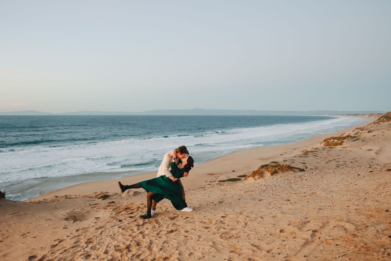 Dancing couple in elegant attire share a passionate dip pose on sandy beach at sunset.