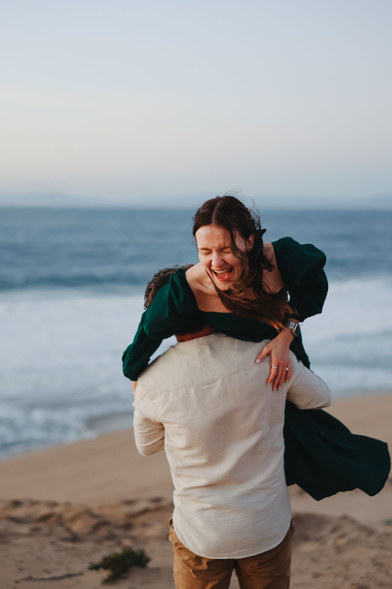 Playful moment captured as person in white shirt lifts partner wearing emerald dress on beach.