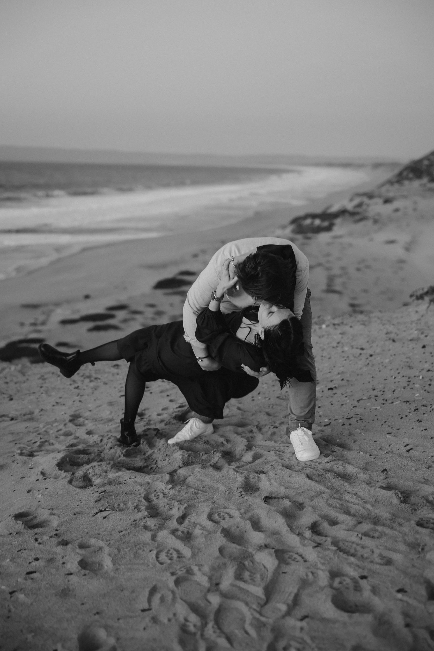 A black and white photo captures a romantic dance pose on a windswept beach.