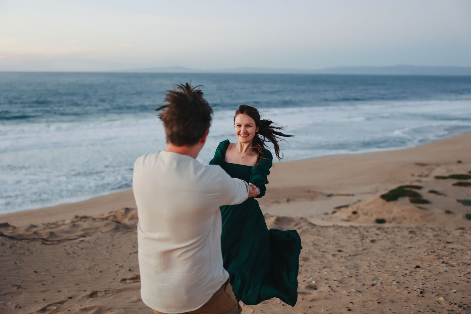 A couple dances playfully on a sandy beach with ocean waves in the background.