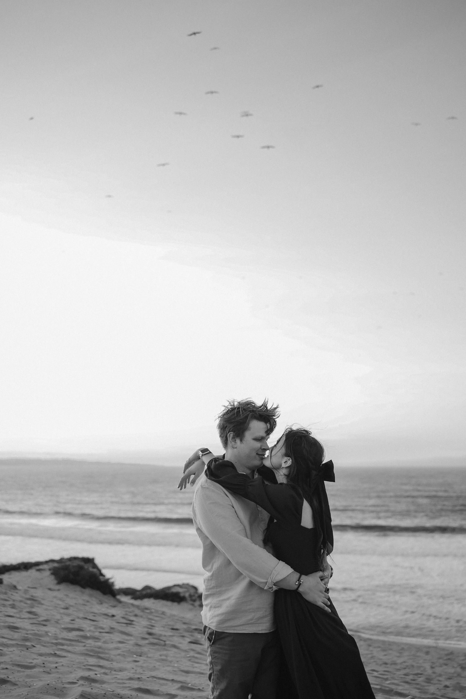 An intimate black and white portrait of a couple embracing on a beach at sunset.