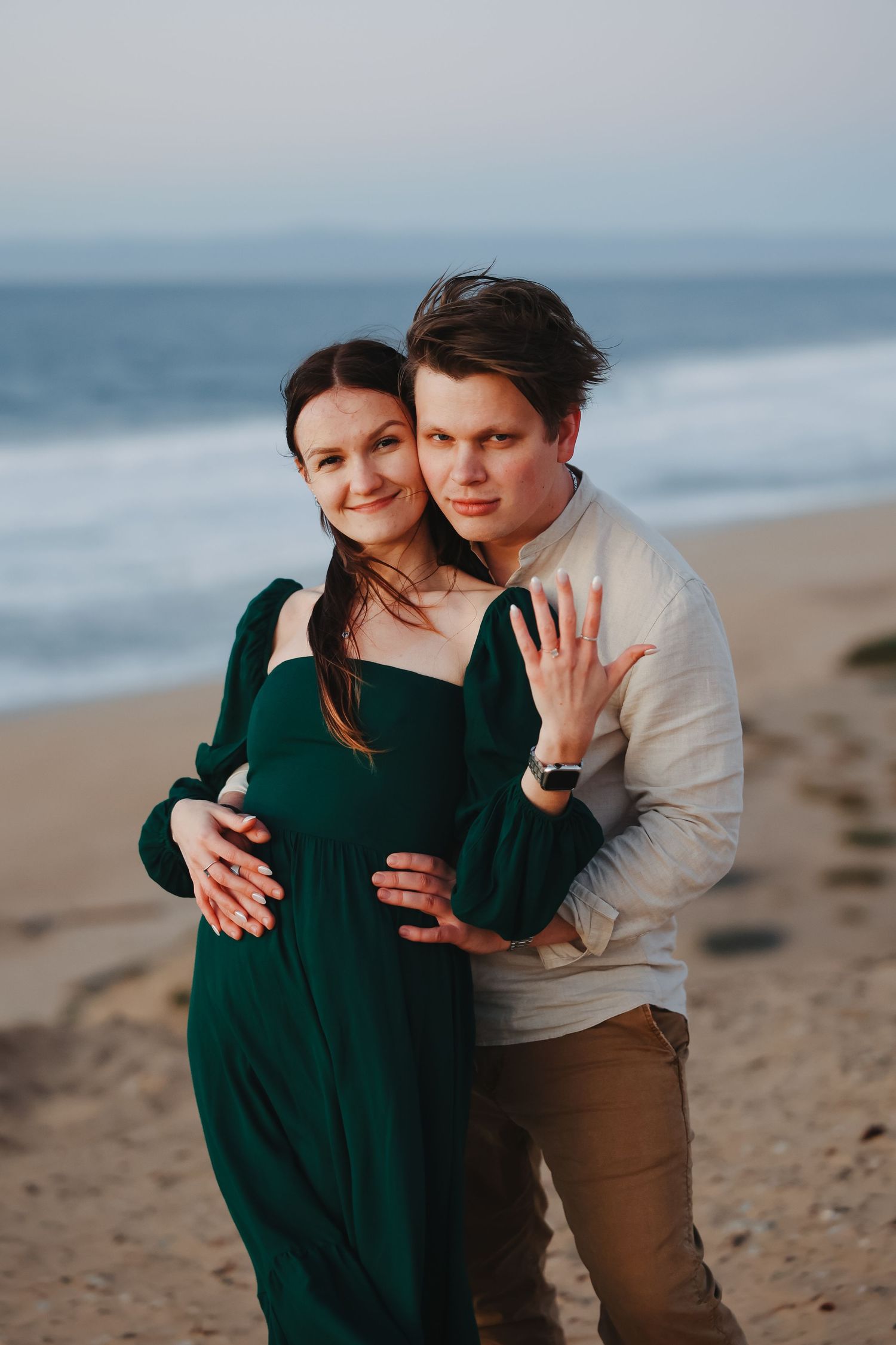 Engagement photo showing a loving embrace on a beach at dusk with a ring prominently displayed.