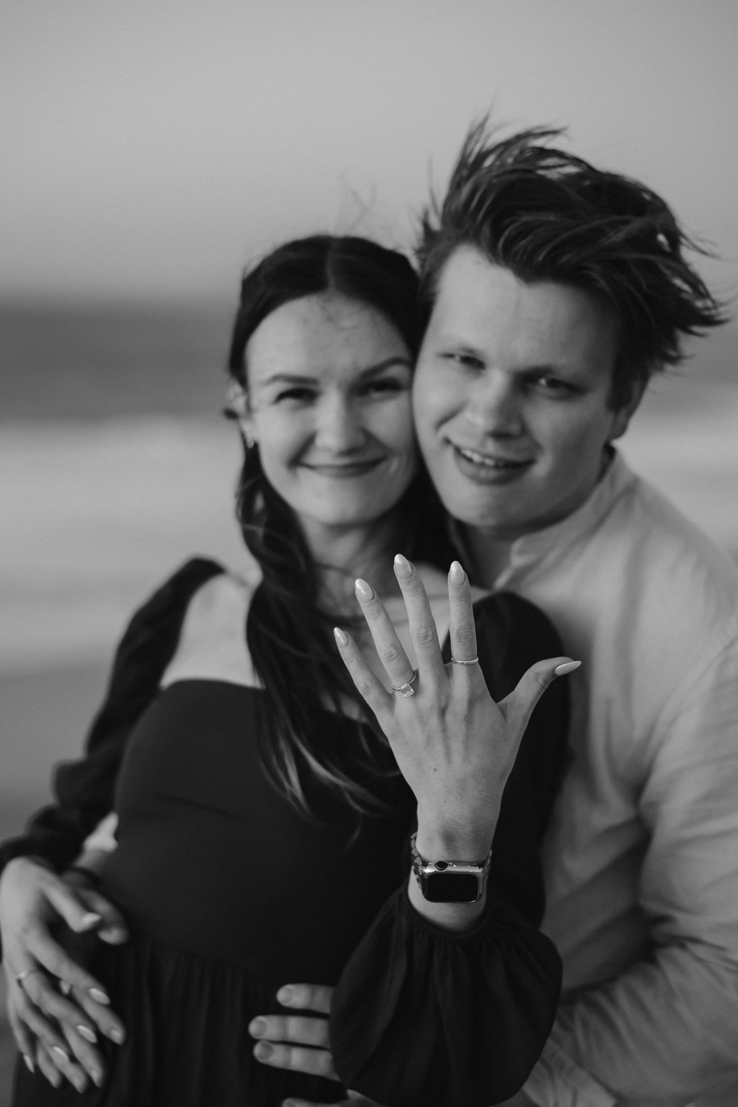Black and white engagement portrait featuring a newly engaged couple displaying the ring on a beach.