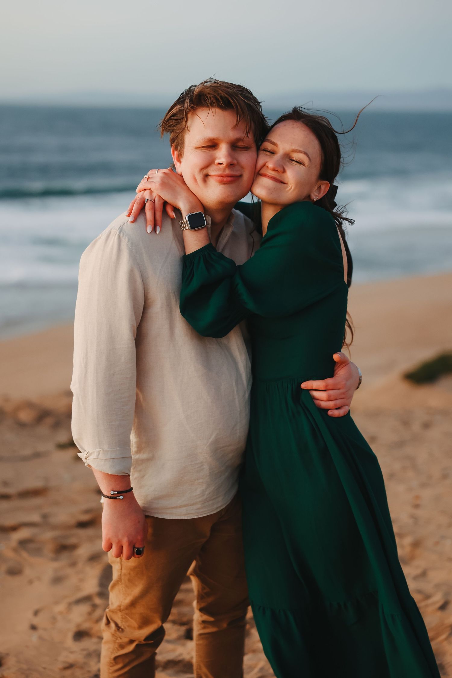 A tender embrace between two people in casual attire standing on a sandy beach with ocean waves in the background.
