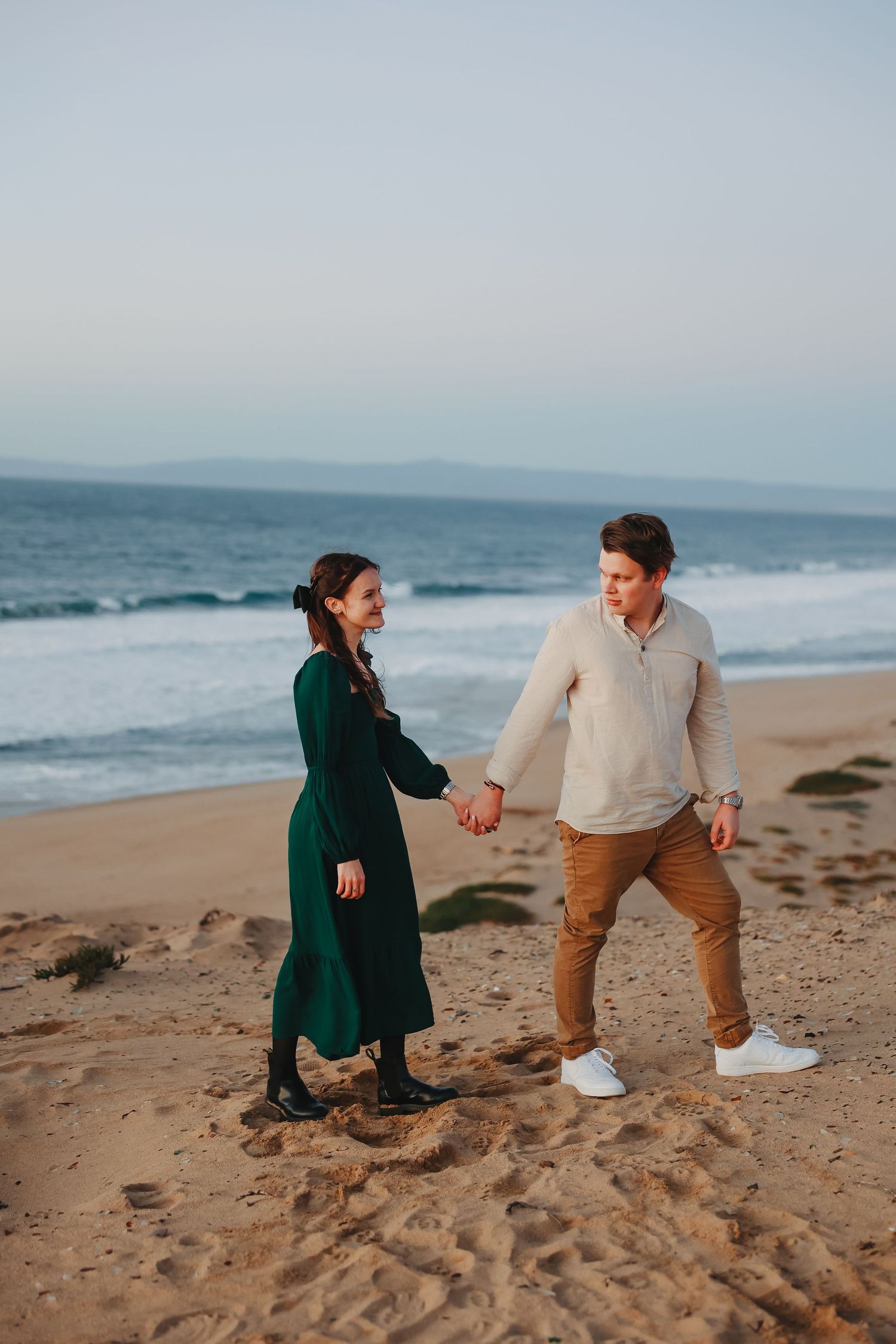 A couple in casual attire holds hands while walking on a beach during golden hour.