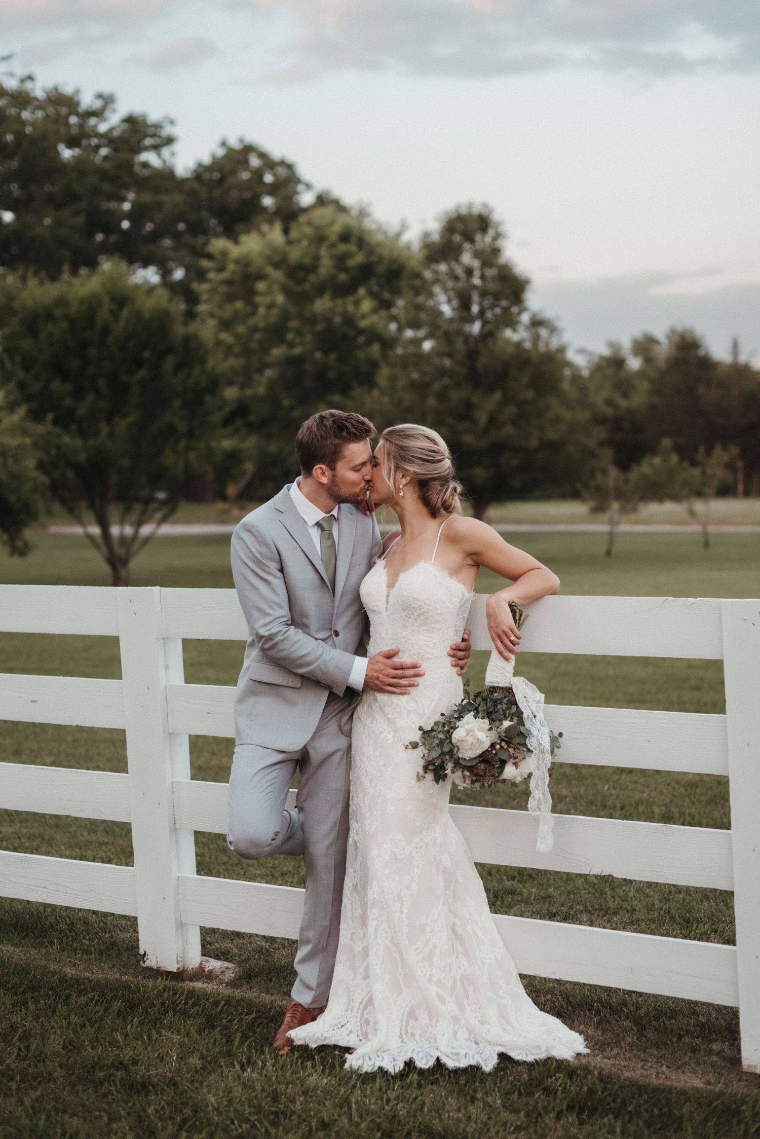 Wedding couple shares intimate moment by white fence during countryside photoshoot.