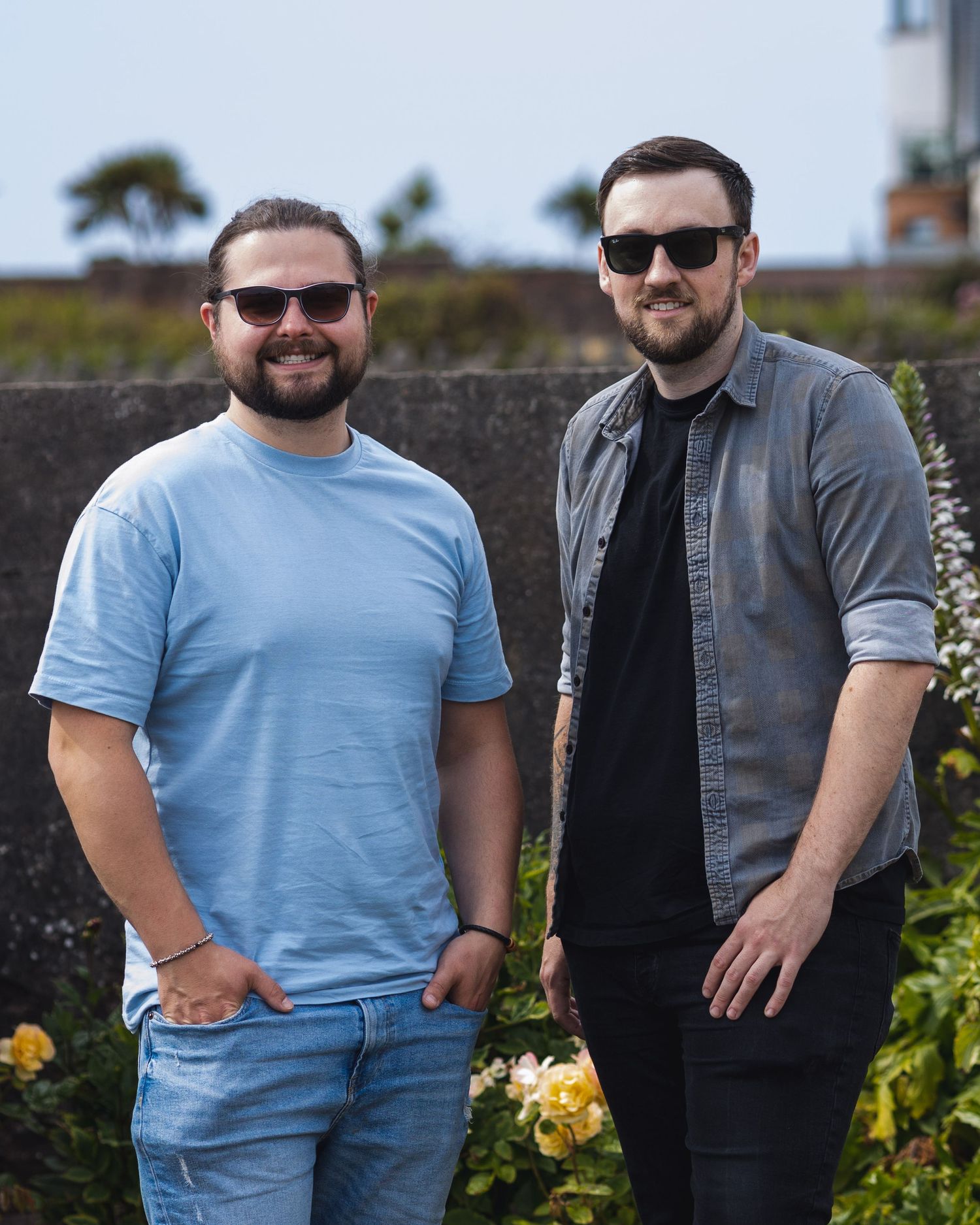 Two people in blue t-shirts and black vests pose together outdoors.