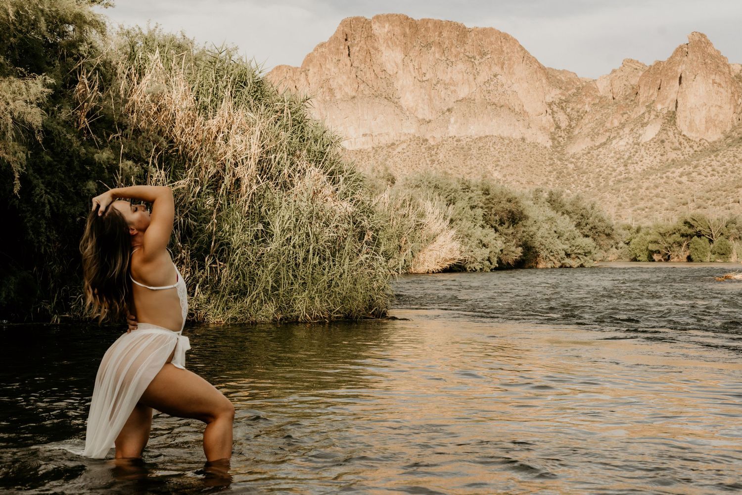 Woman in white dress stands in river with scenic desert mountain landscape.