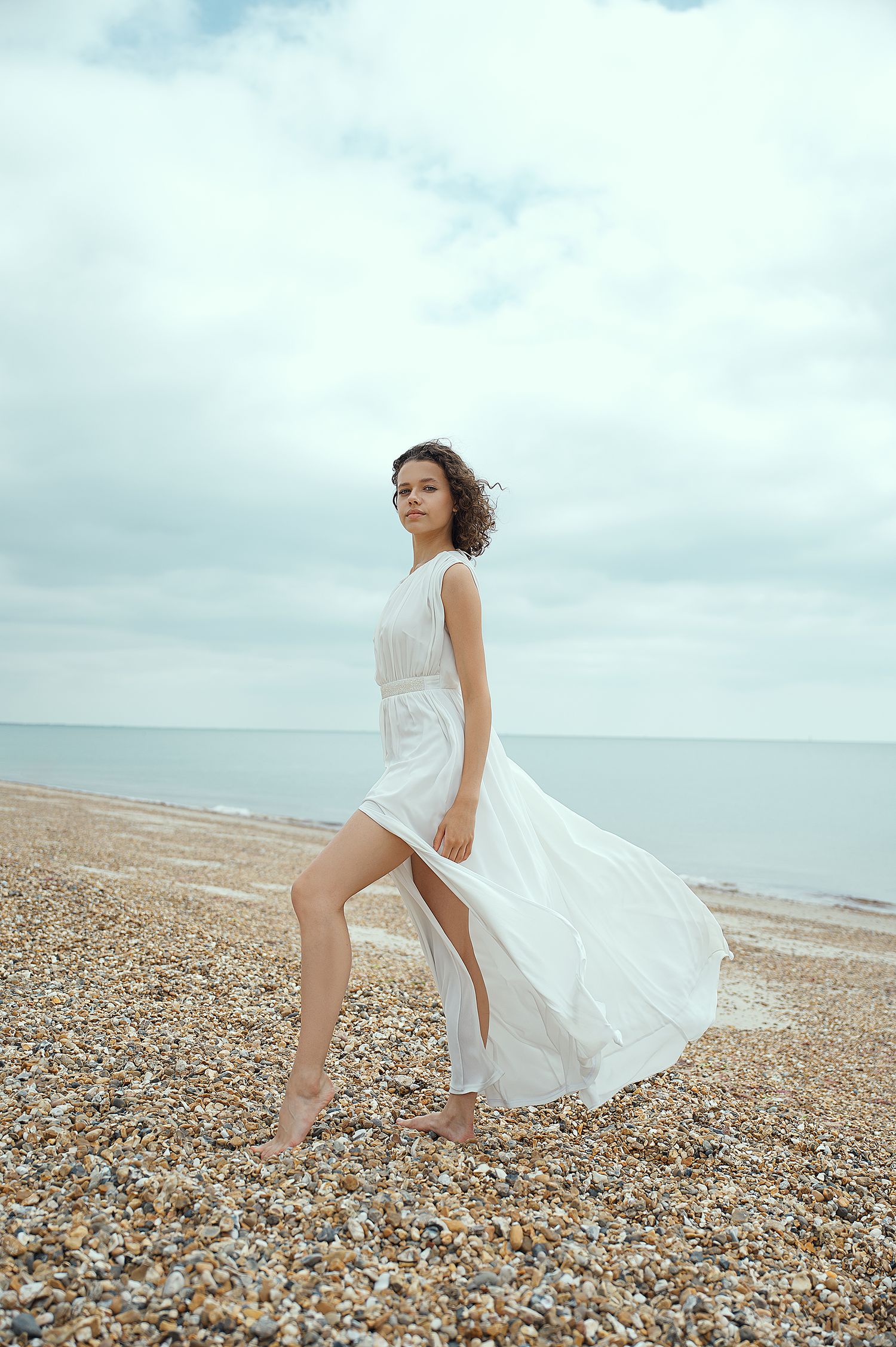 Ethereal white dress flows in beach breeze as woman poses on pebbled shore.