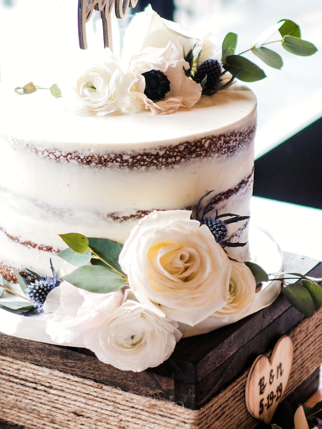 Beautiful semi-naked wedding cake adorned with white roses, black flowers and eucalyptus leaves on a rustic wooden stand.