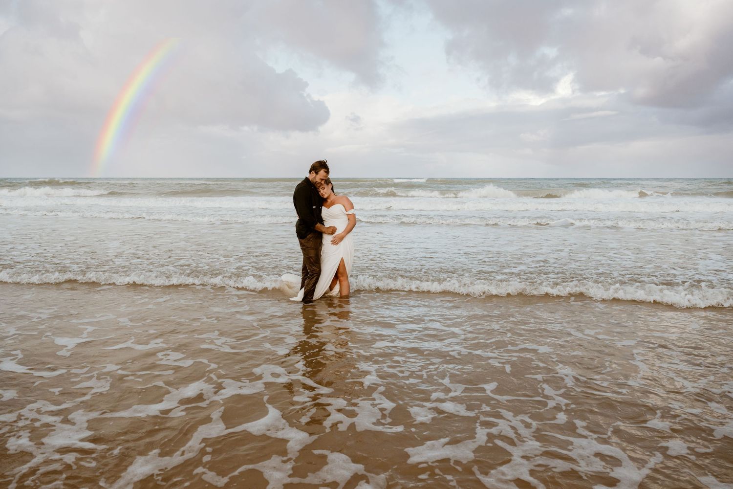 A couple standing in ocean waves with a rainbow arching across the cloudy sky.