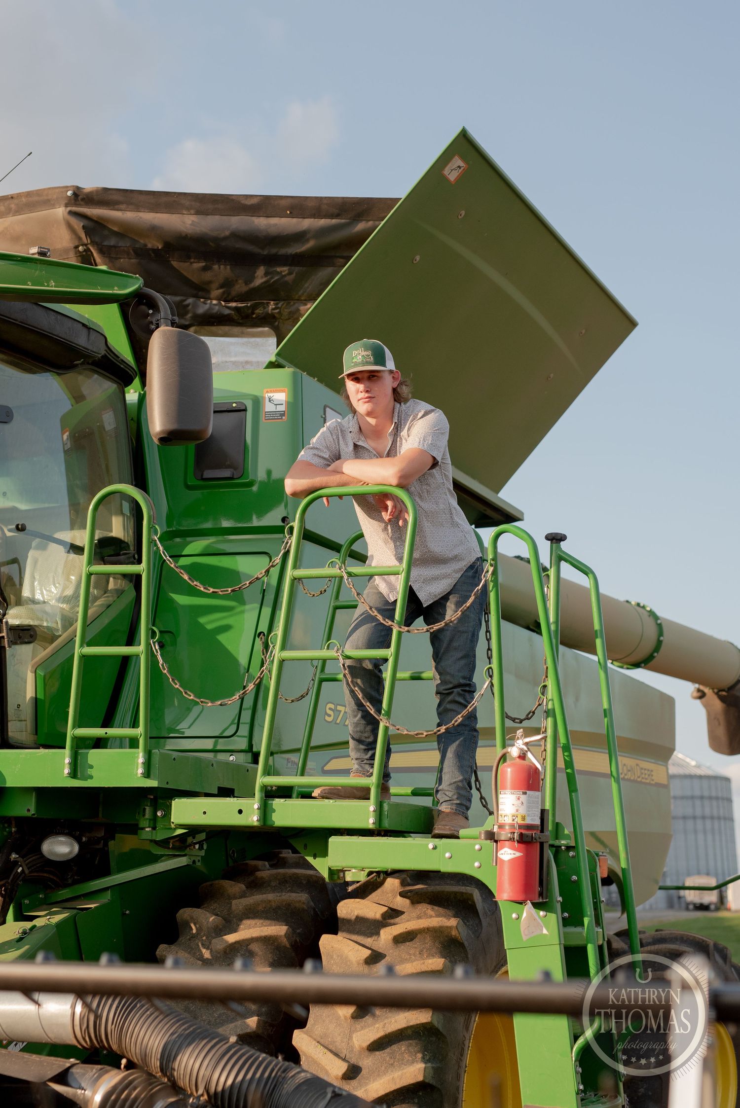 Farmer stands on platform of green combine harvester against blue sky.