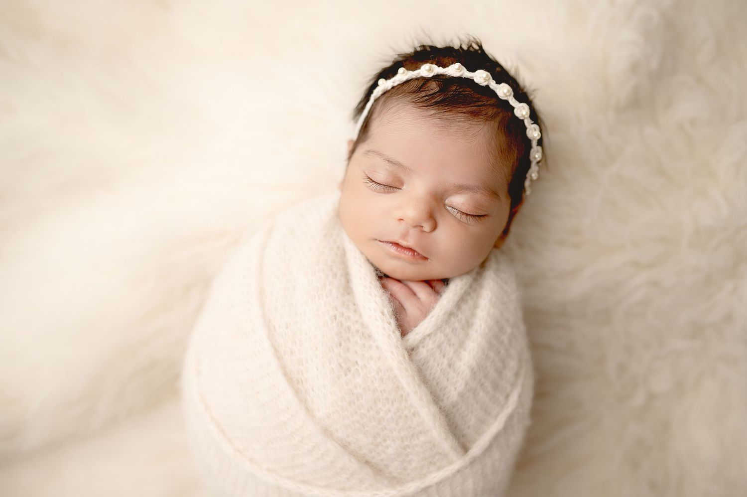 Sleeping newborn wrapped in white blanket wearing delicate headband on cream fur backdrop.