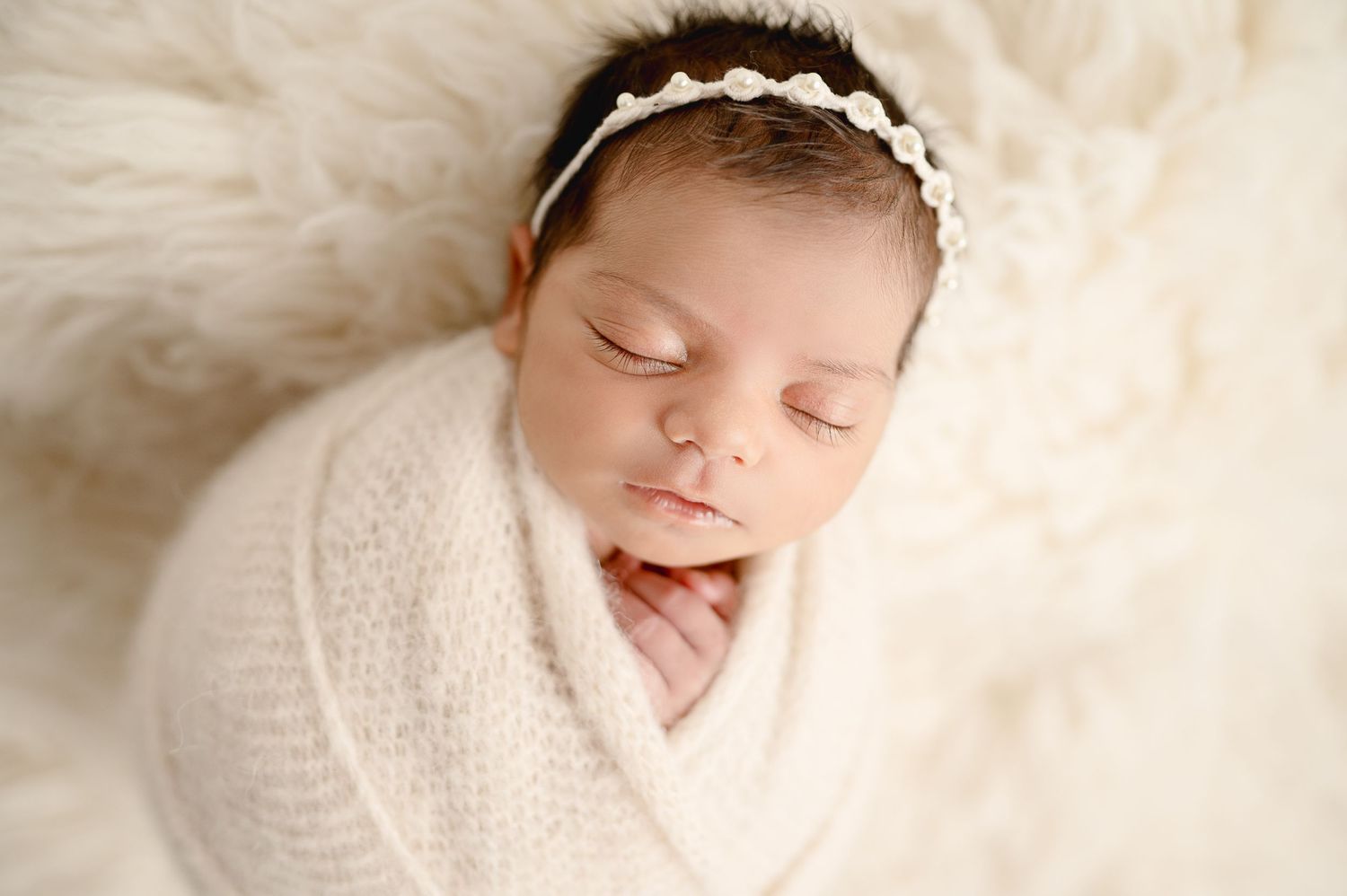 Peaceful newborn baby swaddled in white wrap with lace headband on fluffy cream surface.