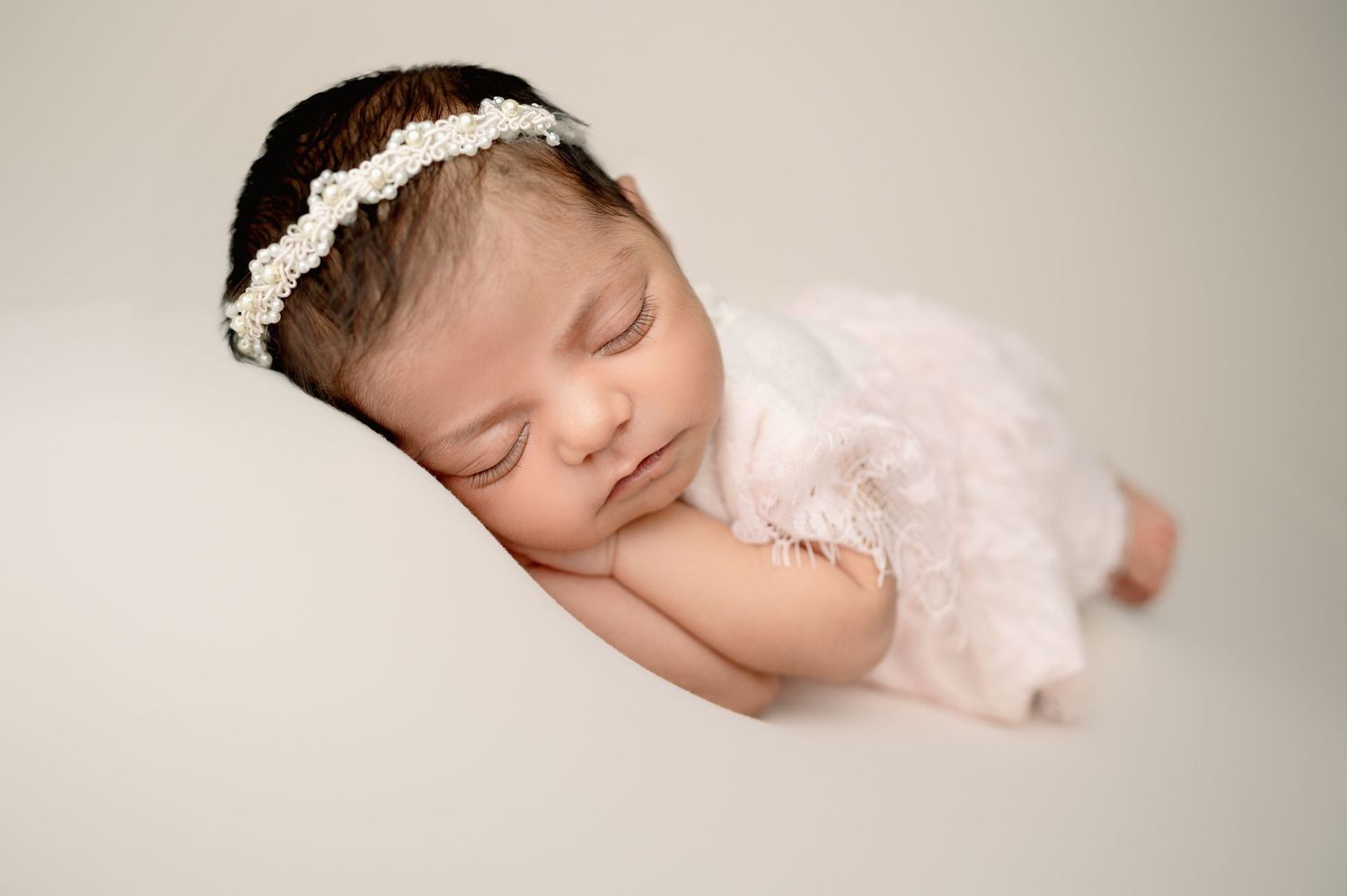 Sleeping baby in soft white lace outfit with headband against neutral backdrop.