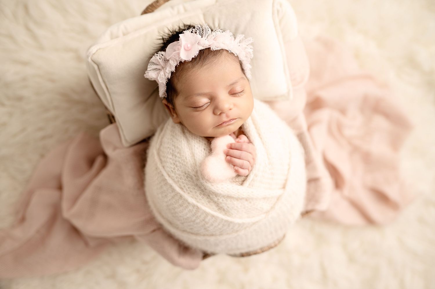 Newborn baby sleeping peacefully in white wrap on pink and cream textured background.