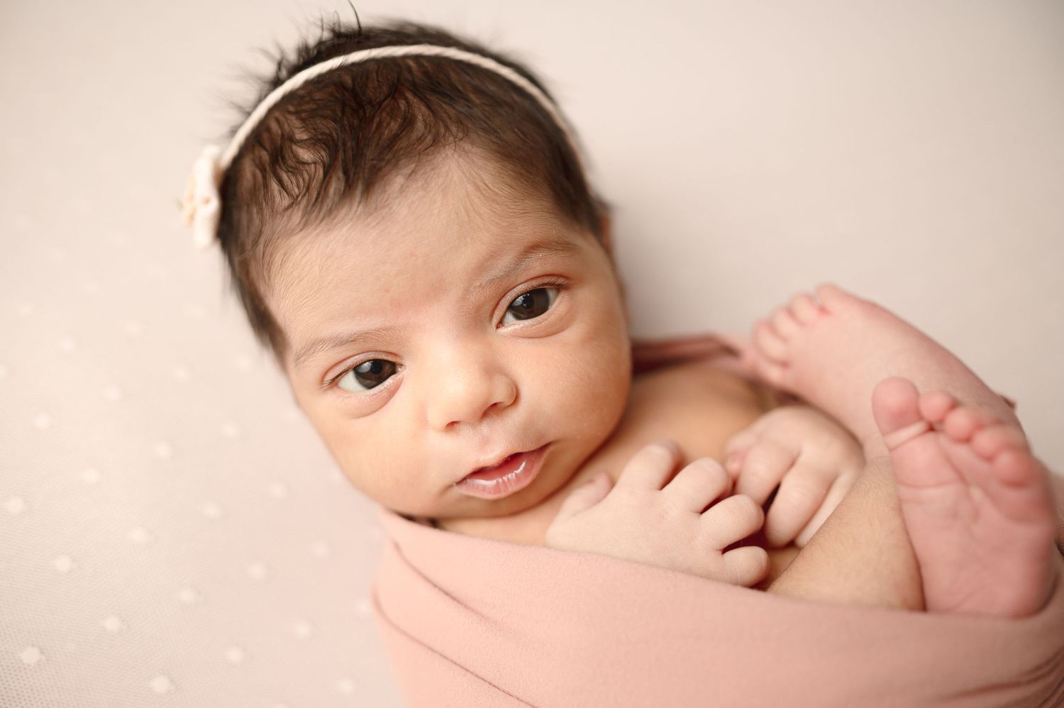 Alert newborn baby wearing headband resting on pink fabric.