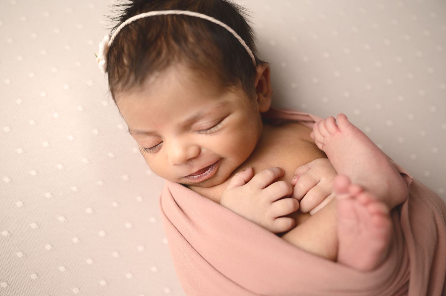 Smiling newborn baby with headband nestled in pink fabric.