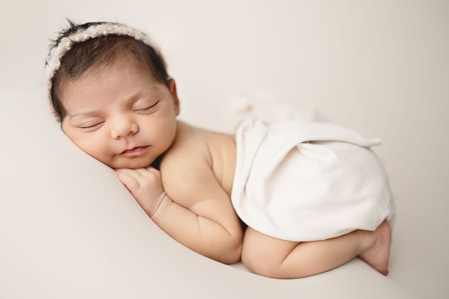 Sleeping newborn in white wrap with delicate headband on neutral background.